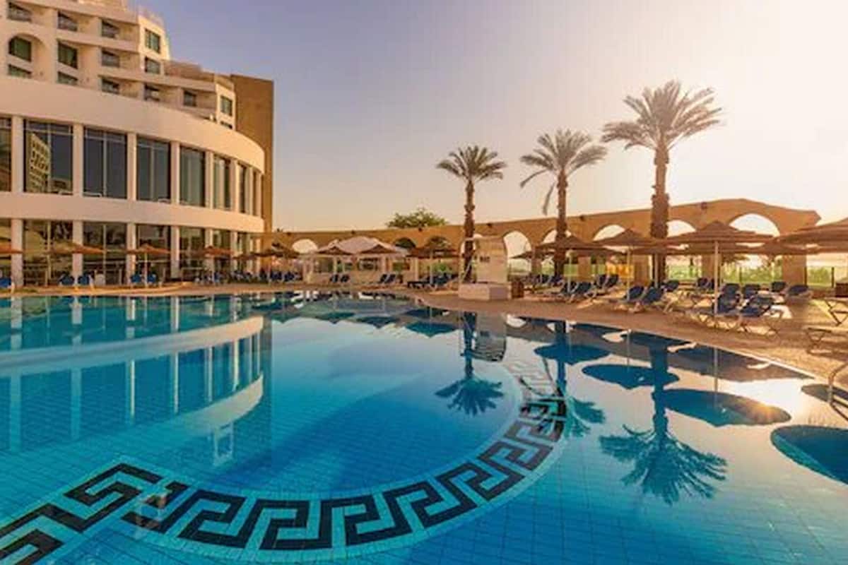 A swimming pool with a geometric tile pattern, surrounded by lounge chairs and palm trees, with a modern building in the background.