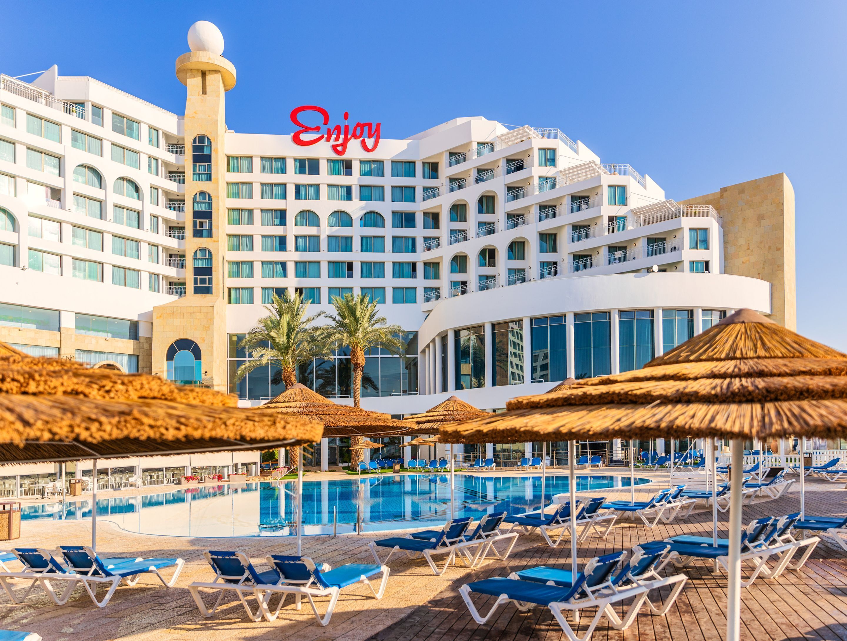A hotel with a swimming pool, sun loungers, and palm trees.