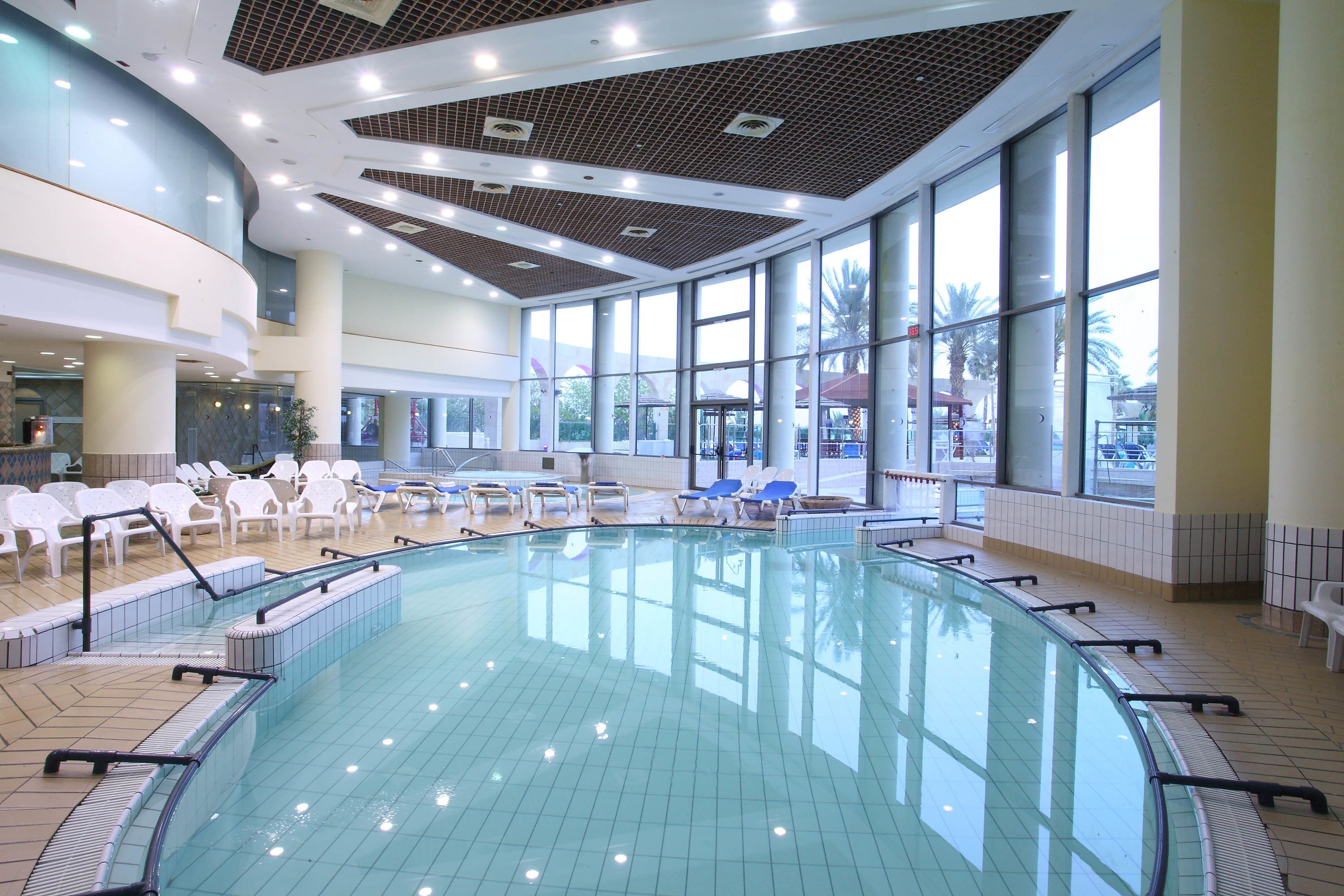 Una amplia piscina interior con paredes de vidrio transparente, rodeada de sillas de descanso y un suelo de baldosas.