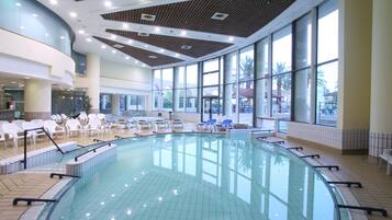 Una amplia piscina interior con paredes de vidrio transparente, rodeada de sillas de descanso y un suelo de baldosas.
