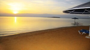 A beach with a clear view of the sunset over the water, a sandy shore, and a structure with a unique roof design. - Brown Dead Sea by Brown Hotels (Tamar)