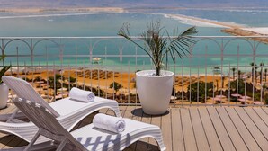 A balcony with two lounge chairs, a potted plant, and a view of a beach and sea. - Brown Dead Sea by Brown Hotels (Tamar)