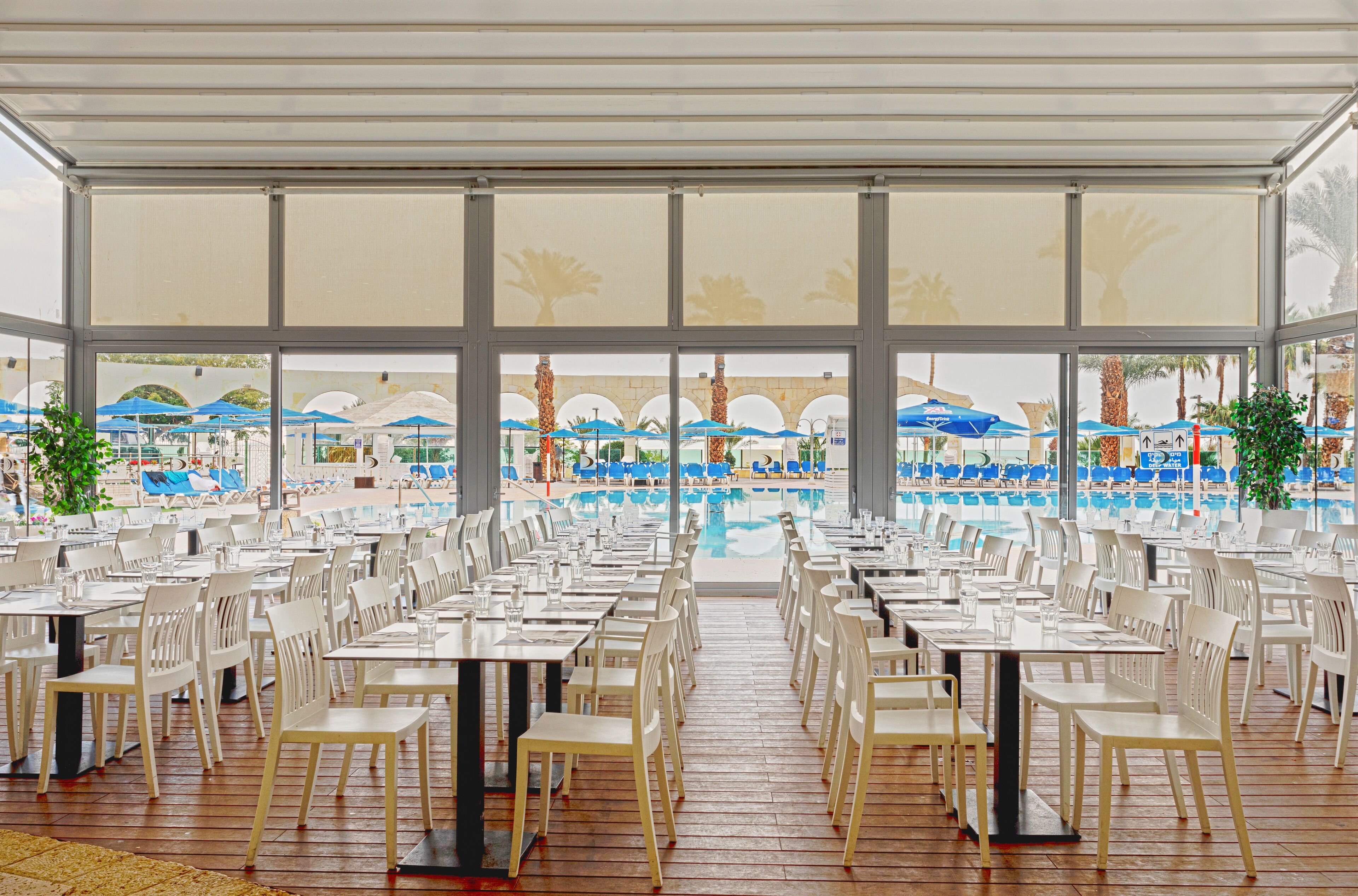 A large indoor pool area with numerous tables and chairs, a view of the pool, and umbrellas in the background.