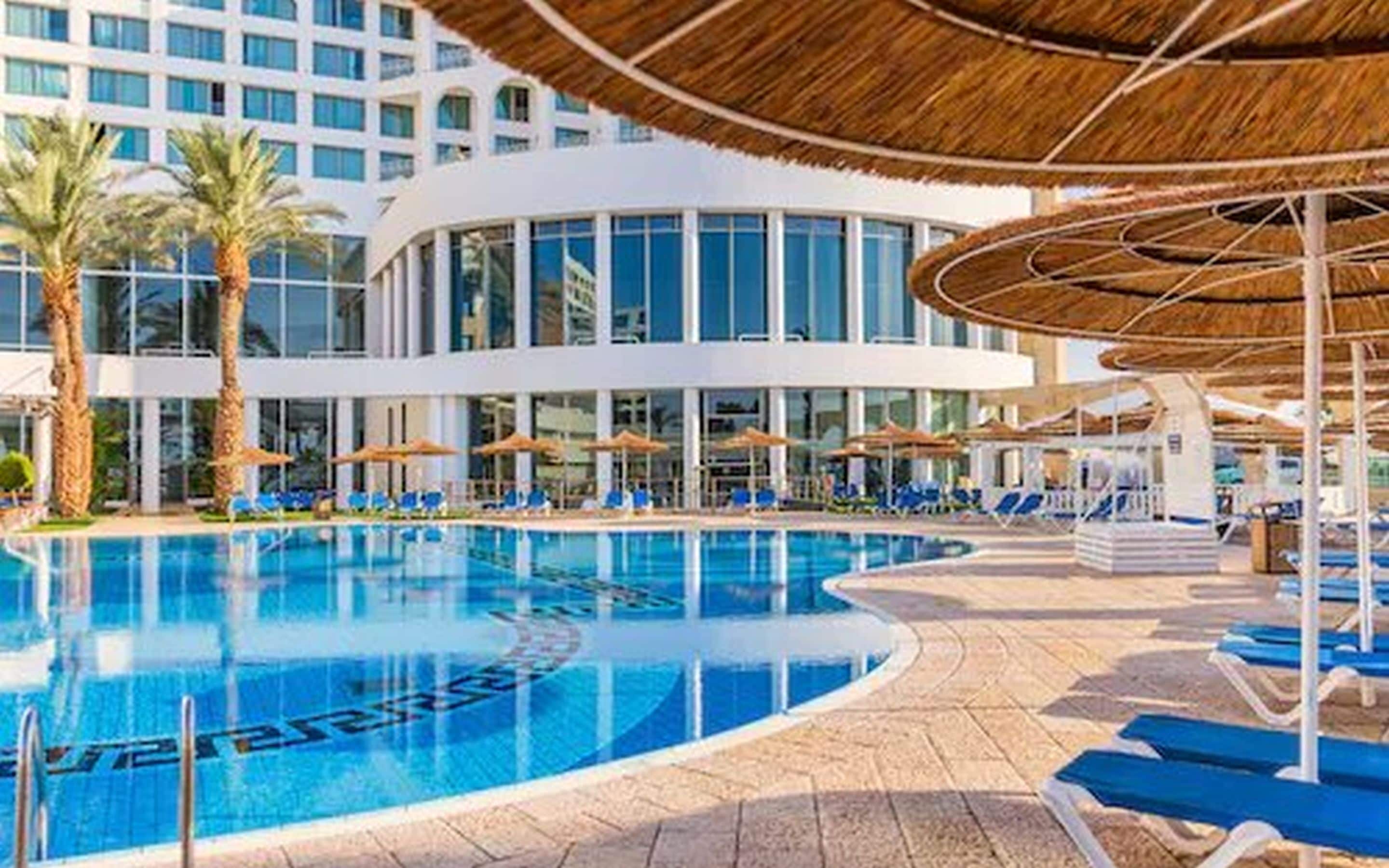 A swimming pool with lounge chairs and umbrellas, surrounded by palm trees and a modern building with large windows.