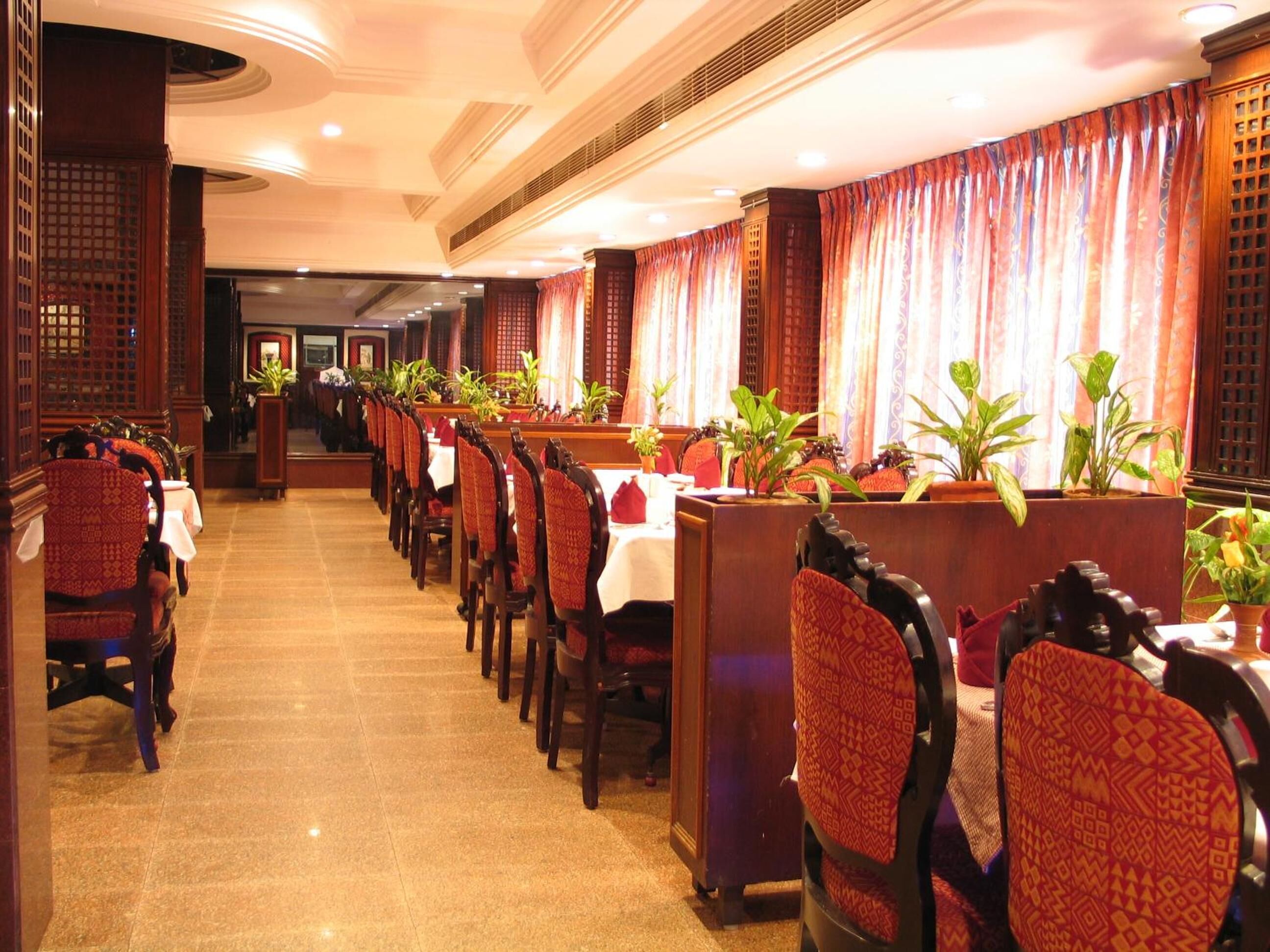 A long corridor with rows of tables and chairs, potted plants, and a traditional architectural style with wooden paneling and a tiled floor.