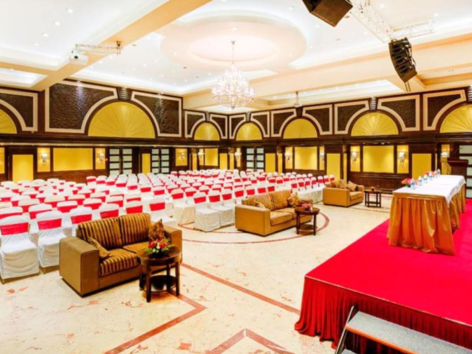 Un gran salón con filas de sillas blancas, un escenario con fondo rojo y una lámpara de araña.