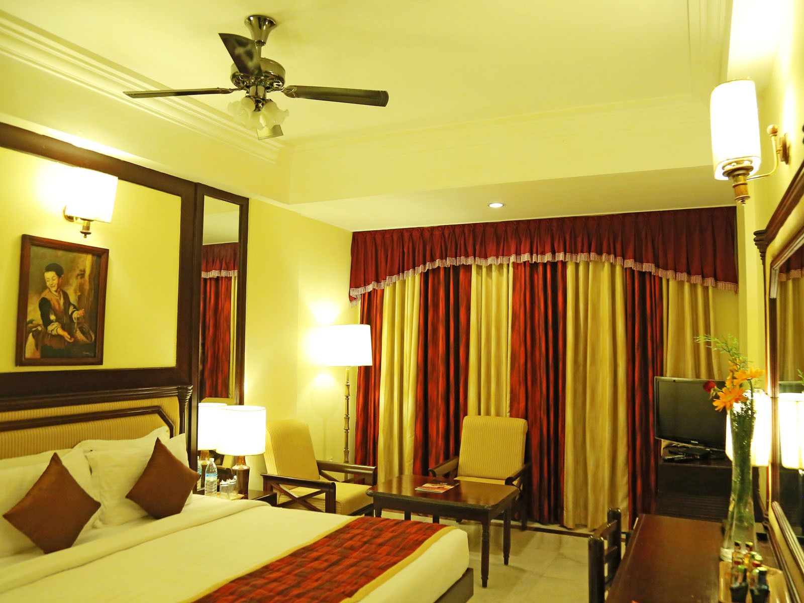 A hotel room with a bed, two chairs, a desk, a television, and a ceiling fan.