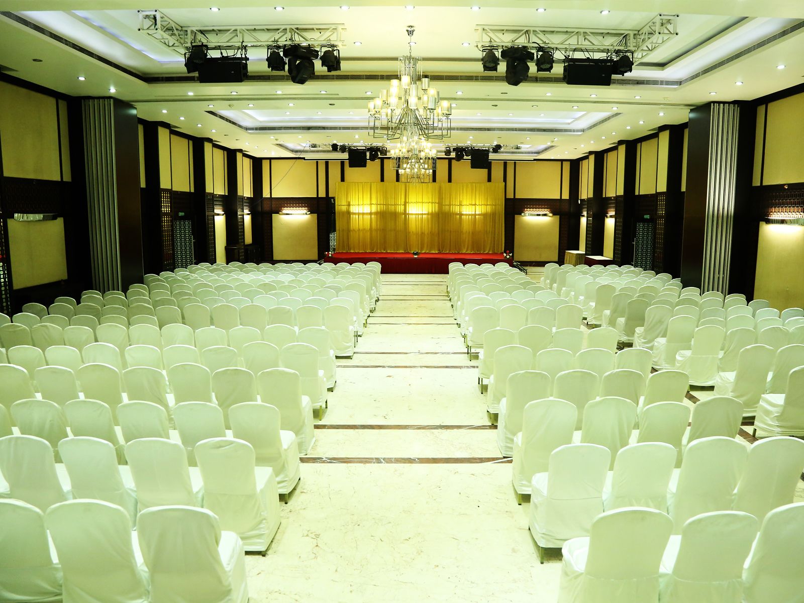 A conference room with rows of white chairs, a stage with a golden curtain, and a chandelier.