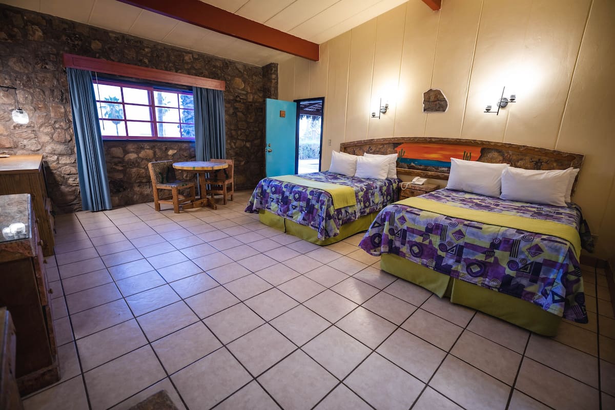 A room with two beds, a wooden desk, a chair, and a stone wall.