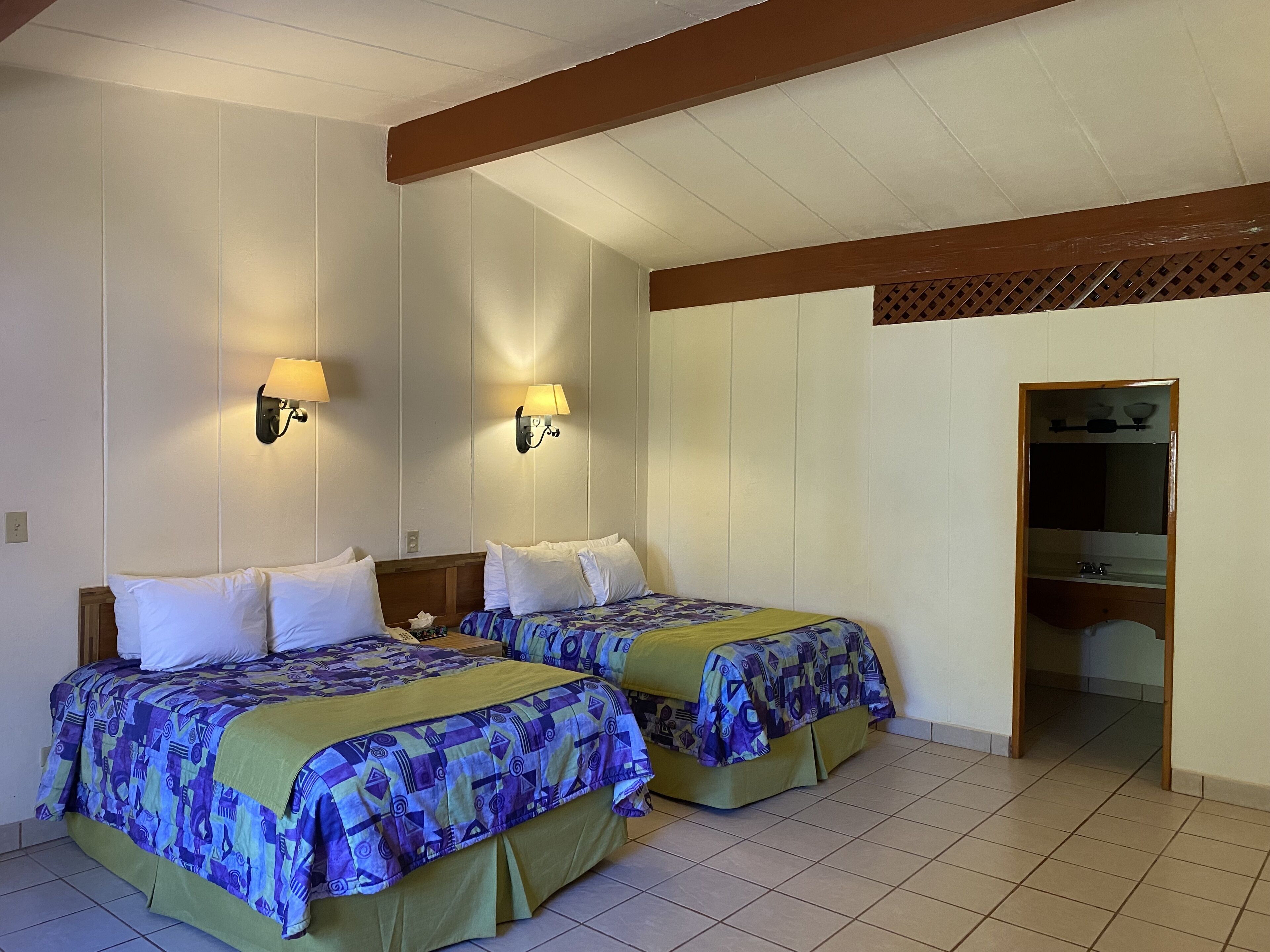 Une chambre d’hôtel avec deux lits, chacun recouvert d’une couvre-lit à motifs, deux lampes fixées au mur et une poutre en bois au plafond.