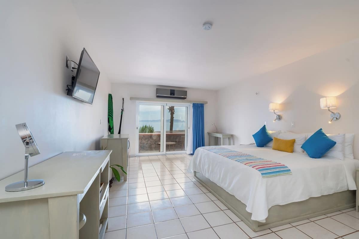 A hotel room with a bed, a desk, a TV, a sliding glass door to a balcony, and a wall-mounted air conditioner.
