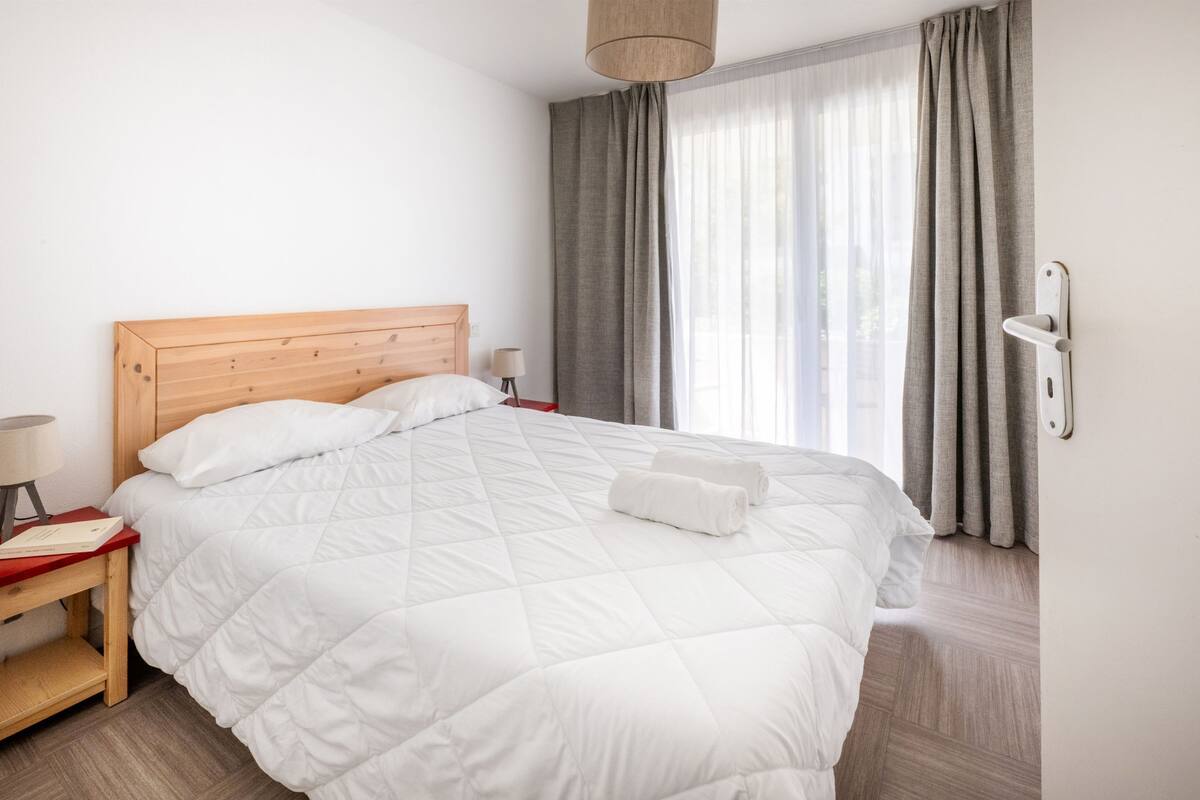 A neatly made bed with white bedding, a wooden headboard, a bedside table with a lamp, and a window with curtains.