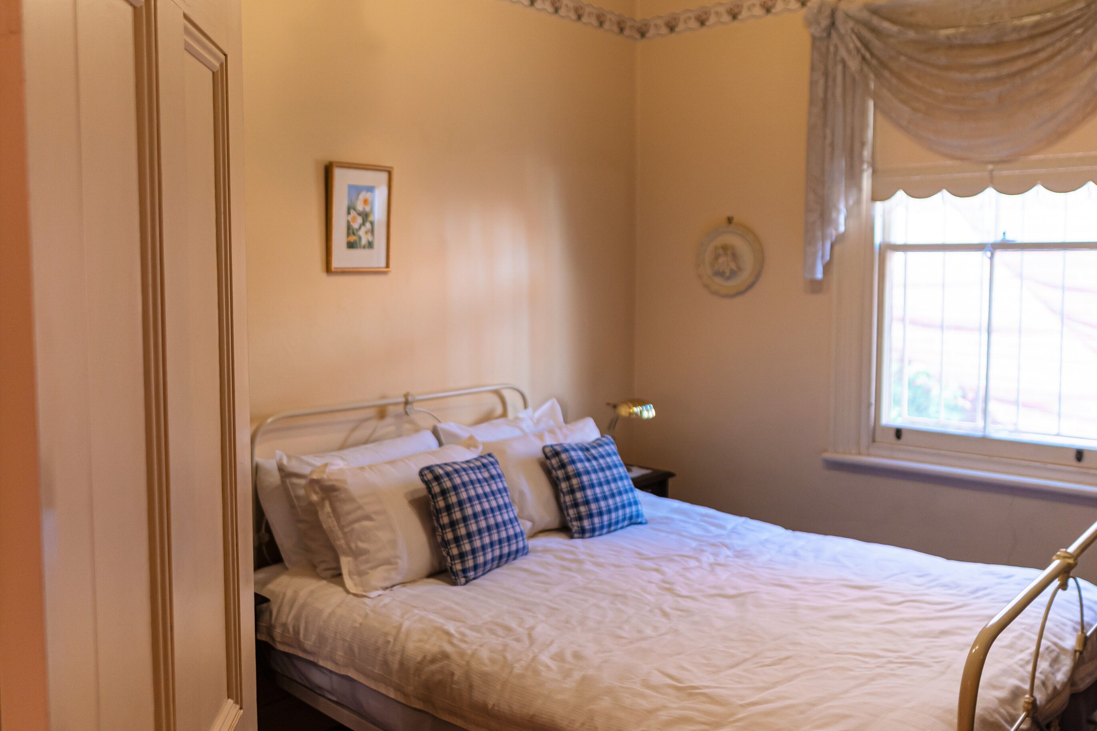 A bedroom with a bed, pillows, a bedside table, a lamp, a window with curtains, and a wall decoration.