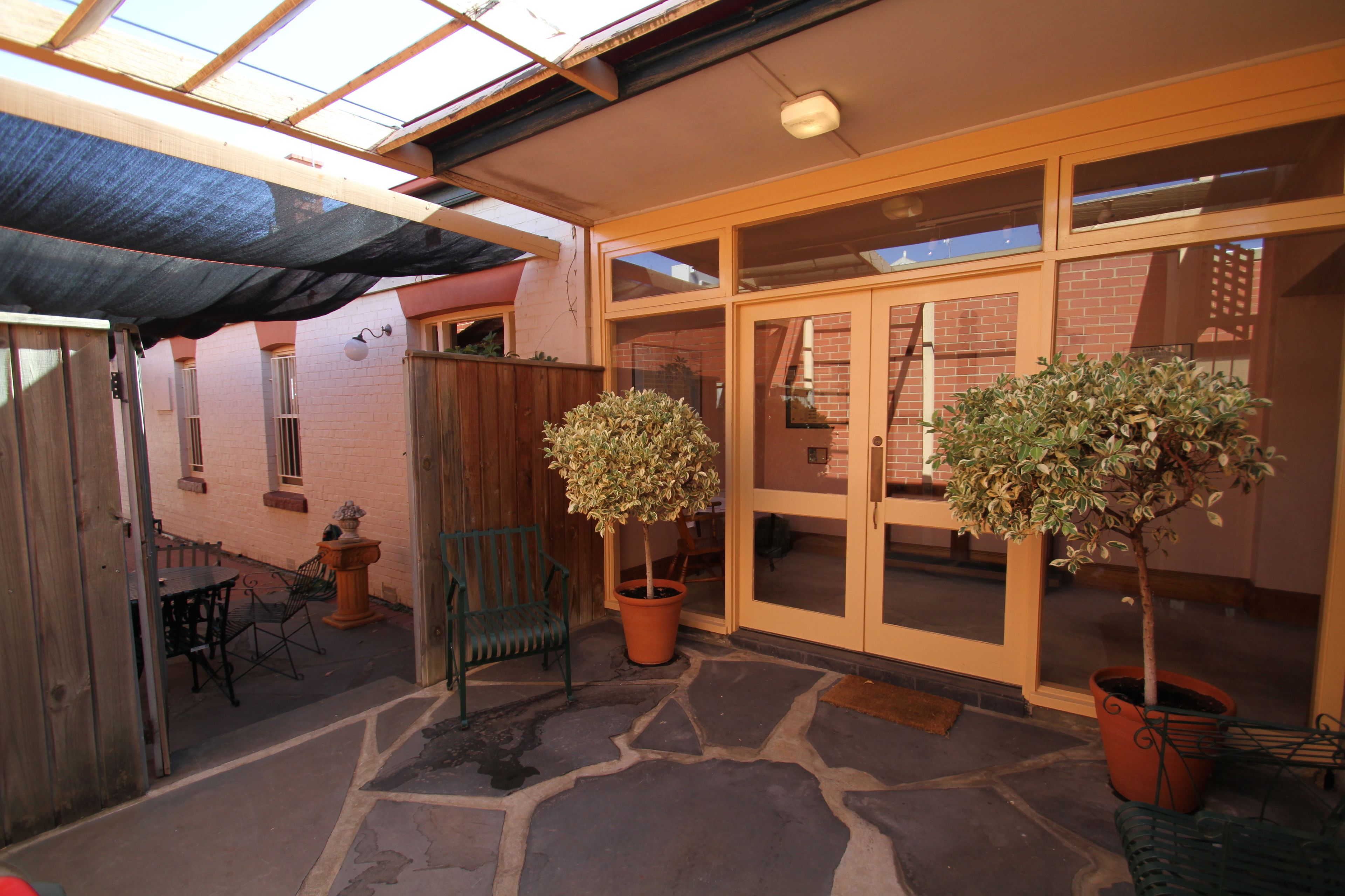 A covered patio area with stone flooring, two potted plants, a wooden fence, and a brick building with glass doors.