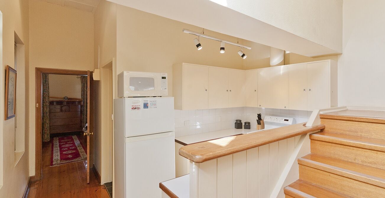 A compact kitchen with a microwave, refrigerator, and wooden countertop. A wooden staircase leads to a landing with a view of a hallway.