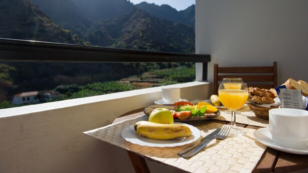 Une table dressée avec des aliments pour le petit-déjeuner, notamment une banane, du jus d’orange et du pain, le tout disposé devant un paysage de montagne.