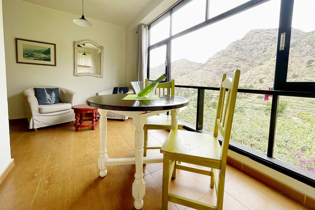 A room with a dining table, chairs, a sofa, and a view of a mountainous landscape through large windows.