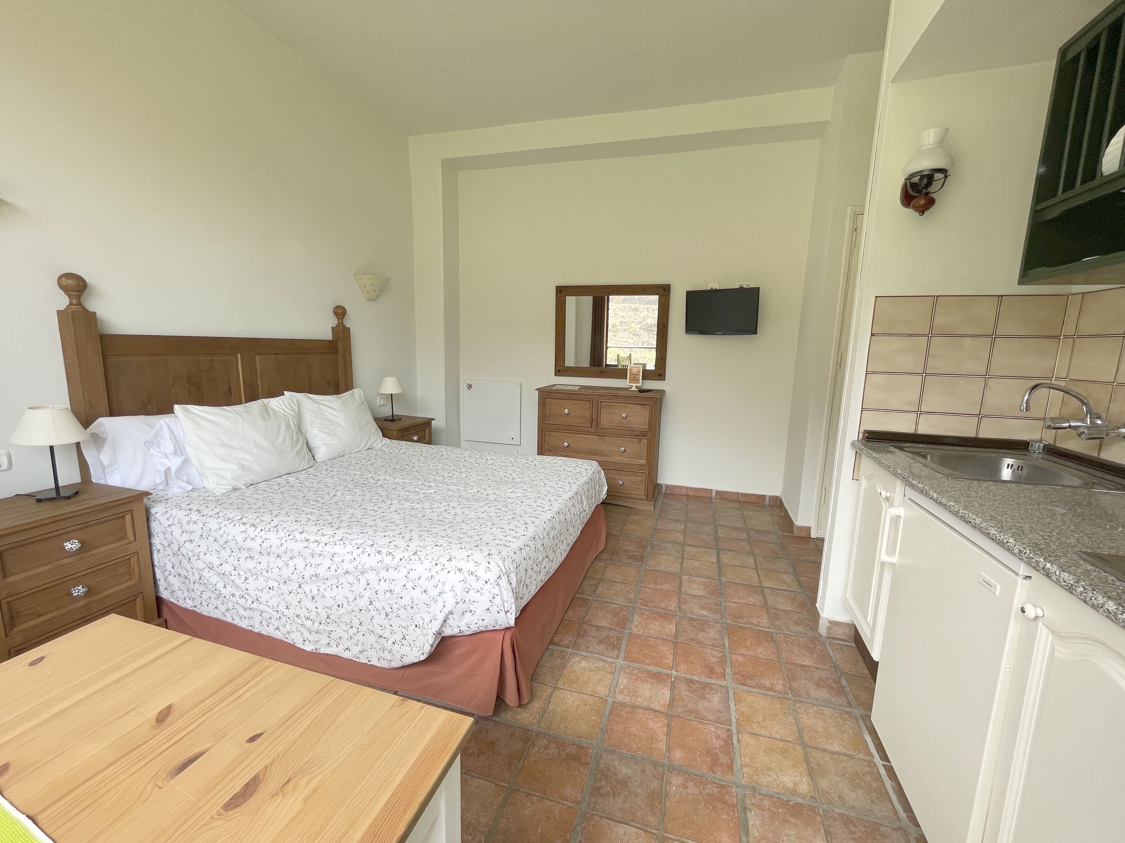 A bedroom with a wooden bed, a bedside table with a lamp, a dresser, and a small mirror.
