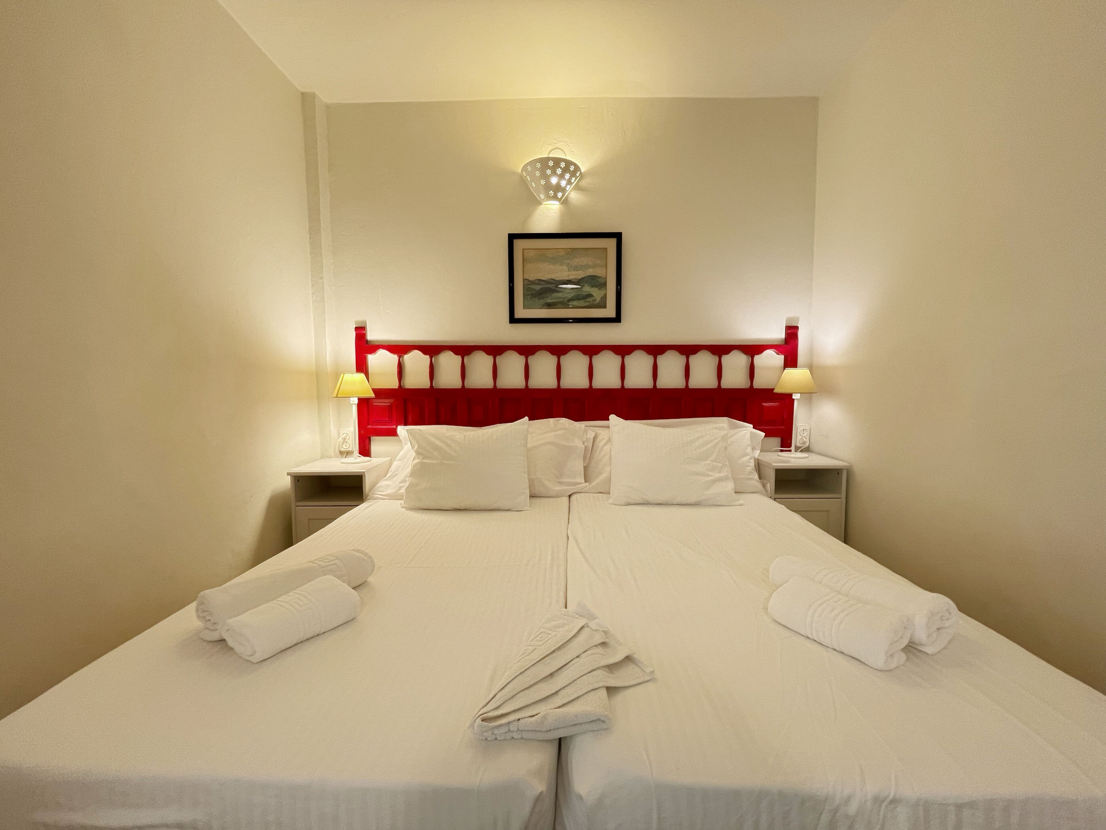 A hotel room with a large bed, two bedside tables, a red headboard, and a framed picture on the wall.