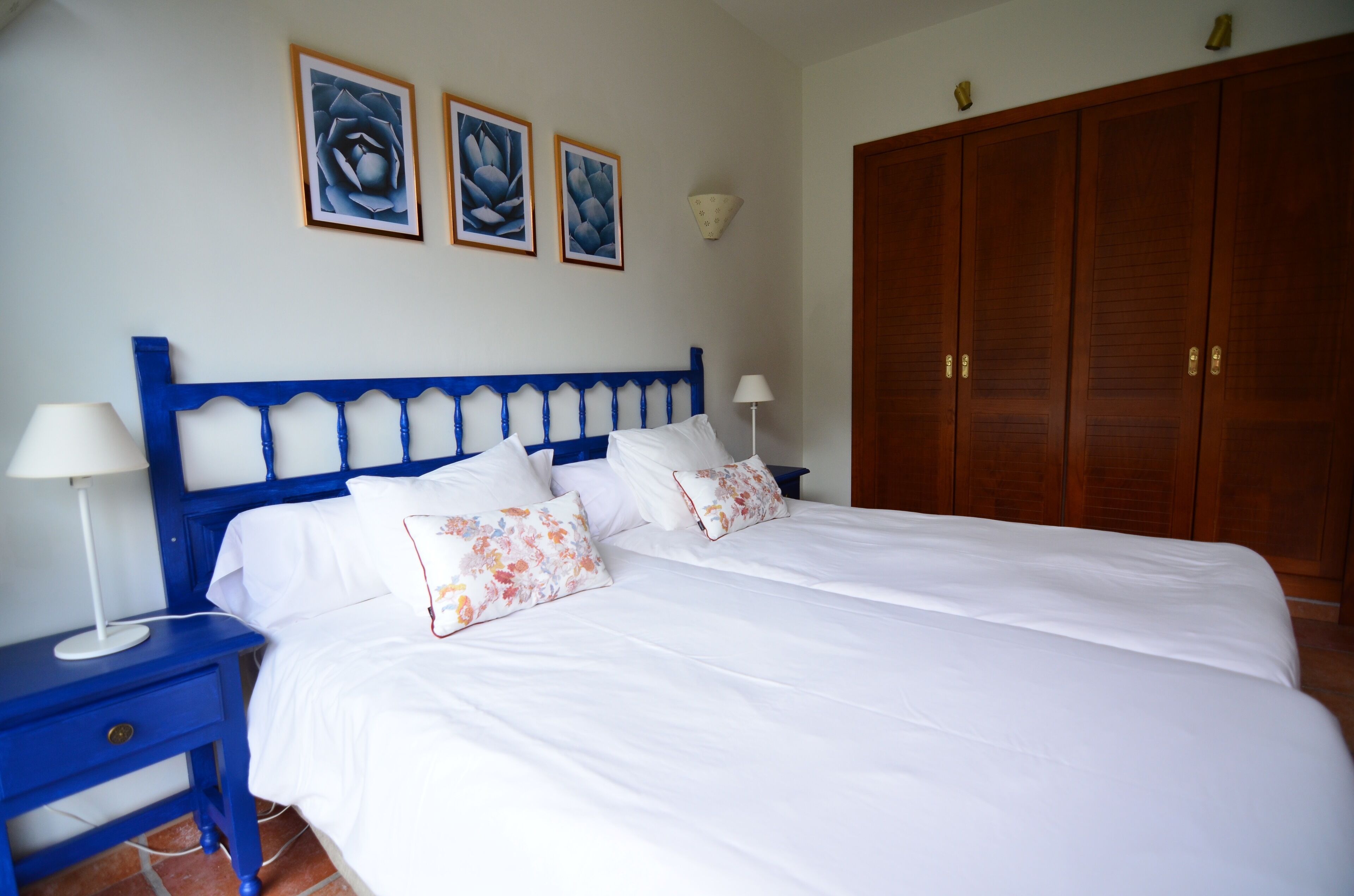 A neatly made bed with white linens and floral pillows, a blue headboard, and a nightstand with a lamp.