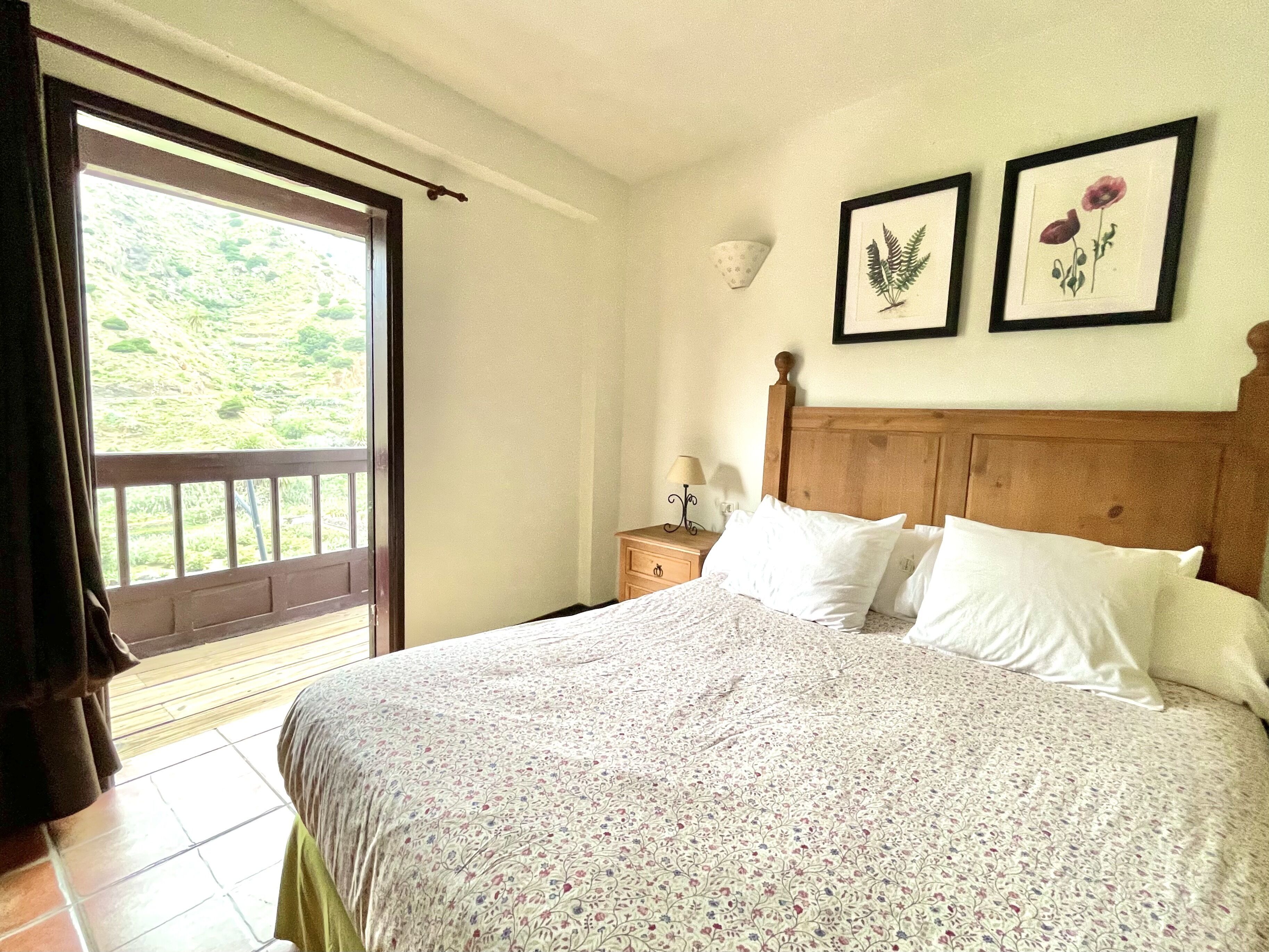 Un dormitorio con cama, mesita de noche, ventana con vista a follaje, y dos láminas botánicas enmarcadas en la pared.