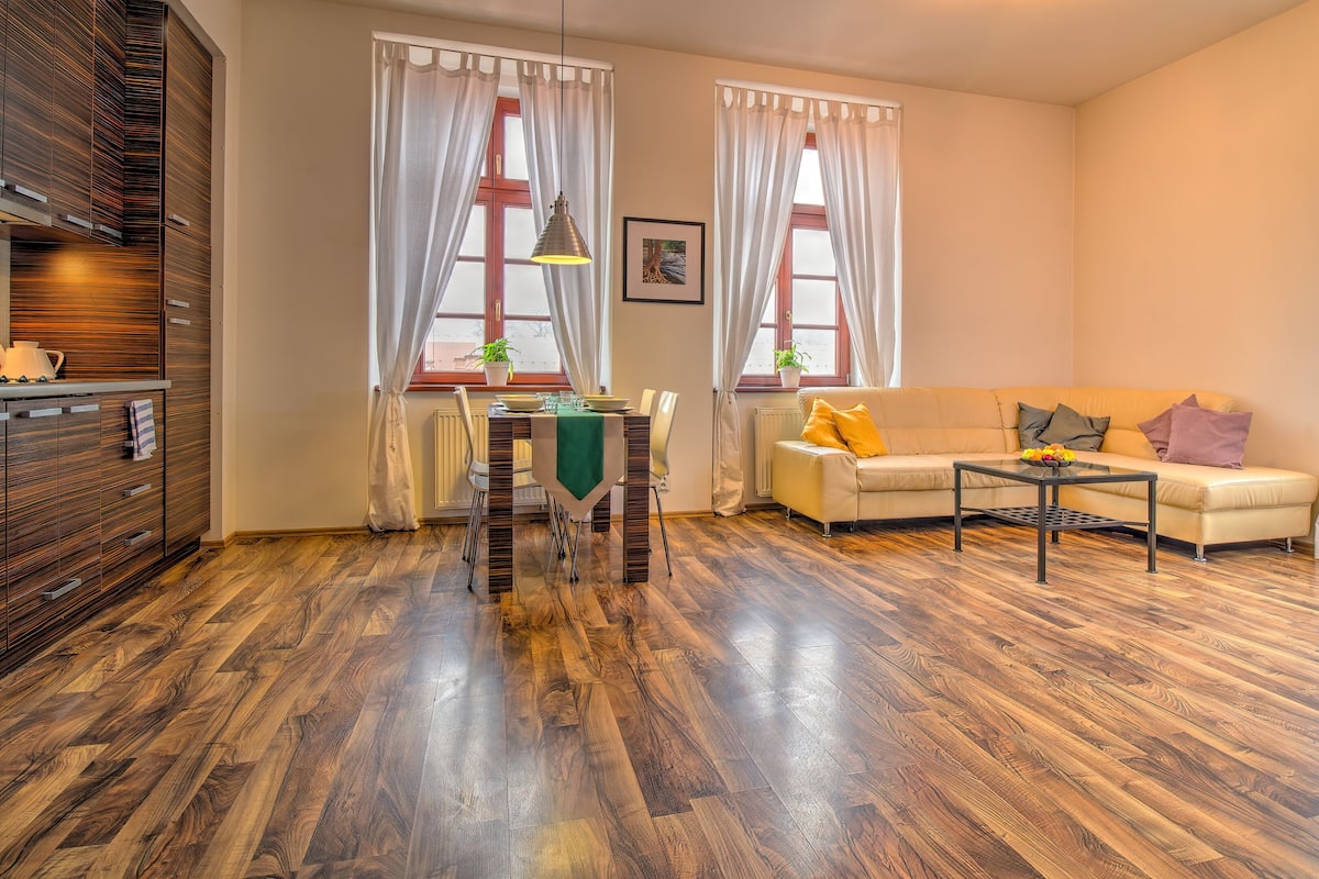 A modern living room with a wooden floor, a sofa, a dining table with chairs, and large windows with curtains.