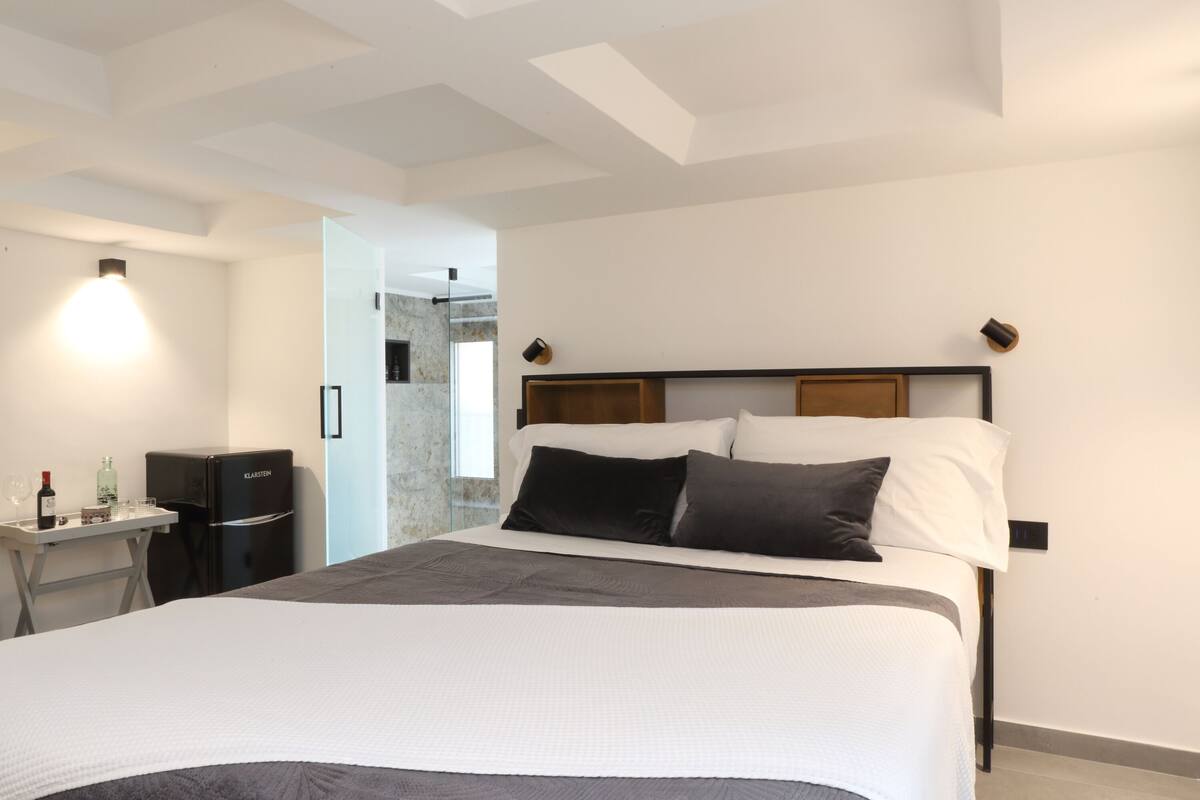 Habitación de hotel moderna con una cama grande, un escritorio pequeño con refrigerador y vista a un muro de piedra a través de una puerta abierta.