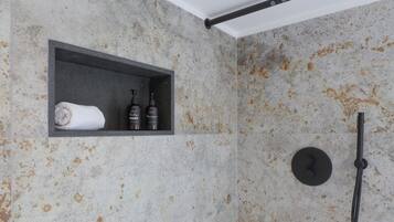 Un baño con pared de mármol, repisa con artículos de aseo y ducha.