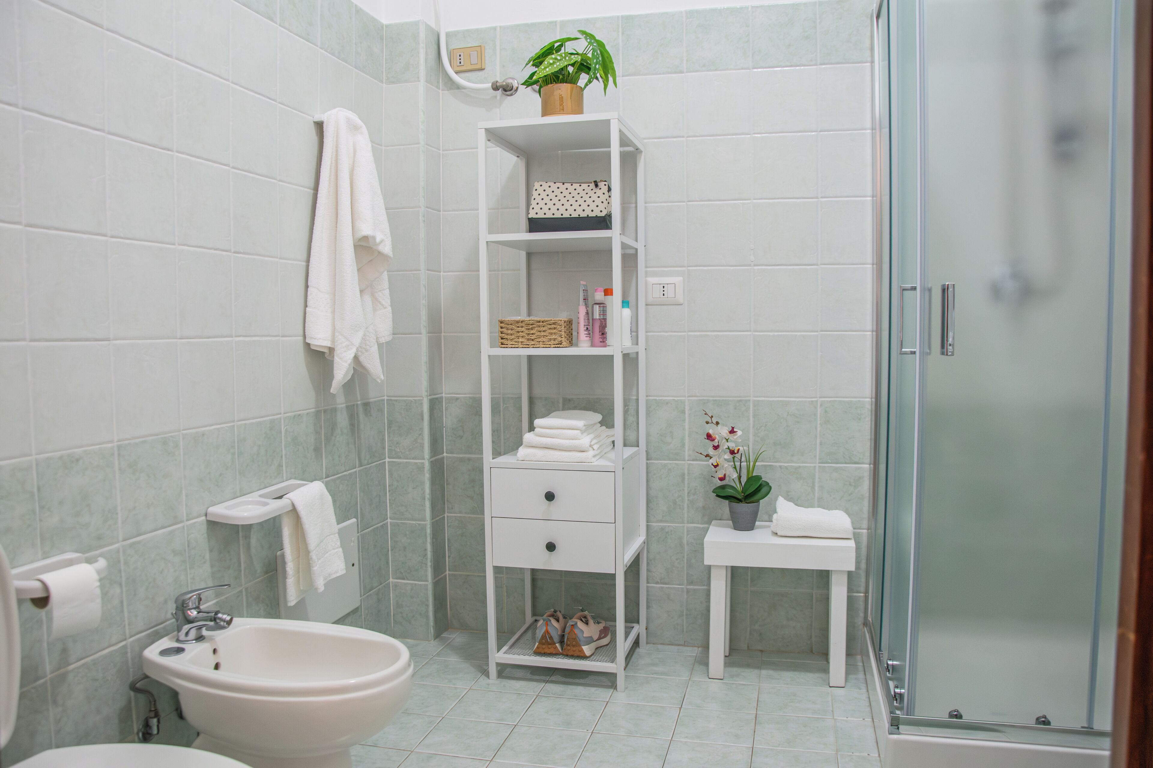 A bathroom with a bidet, a sink, a white shelf with towels and toiletries, and a shower area.