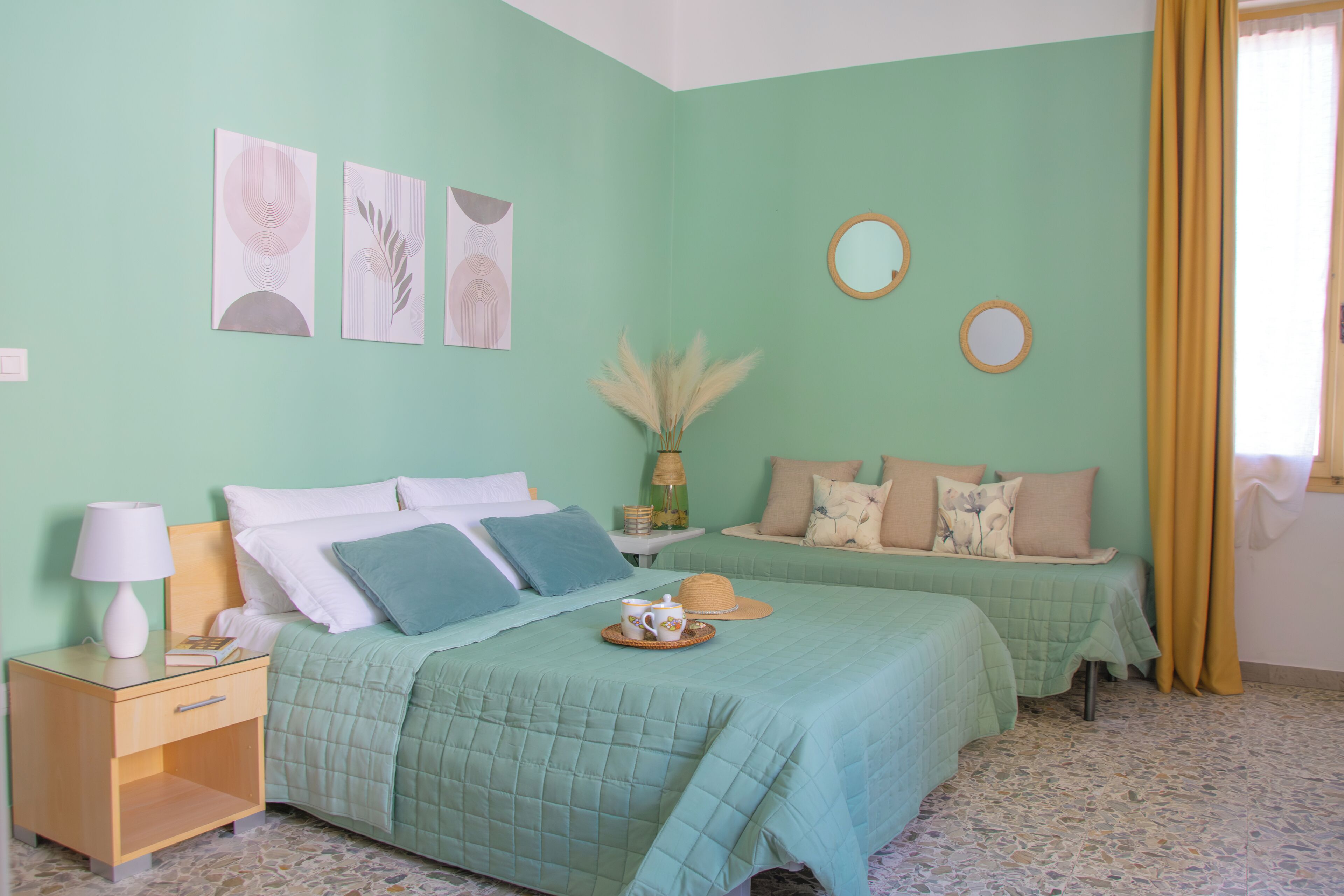 A room with two beds, a nightstand, a lamp, a vase with pampas grass, and two wall mirrors.