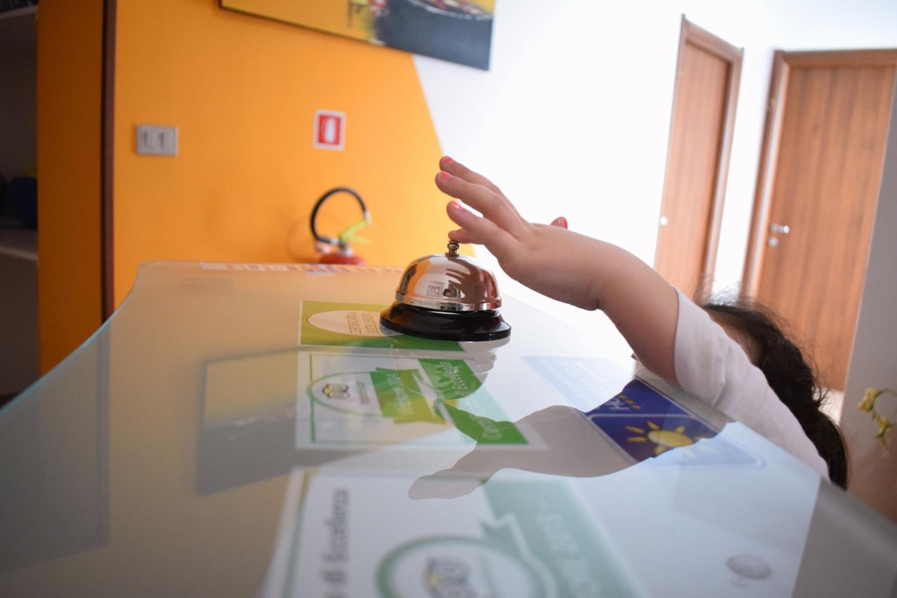 Une main tendue vers une cloche posée sur un comptoir d’accueil, à côté de diverses cartes d’information.