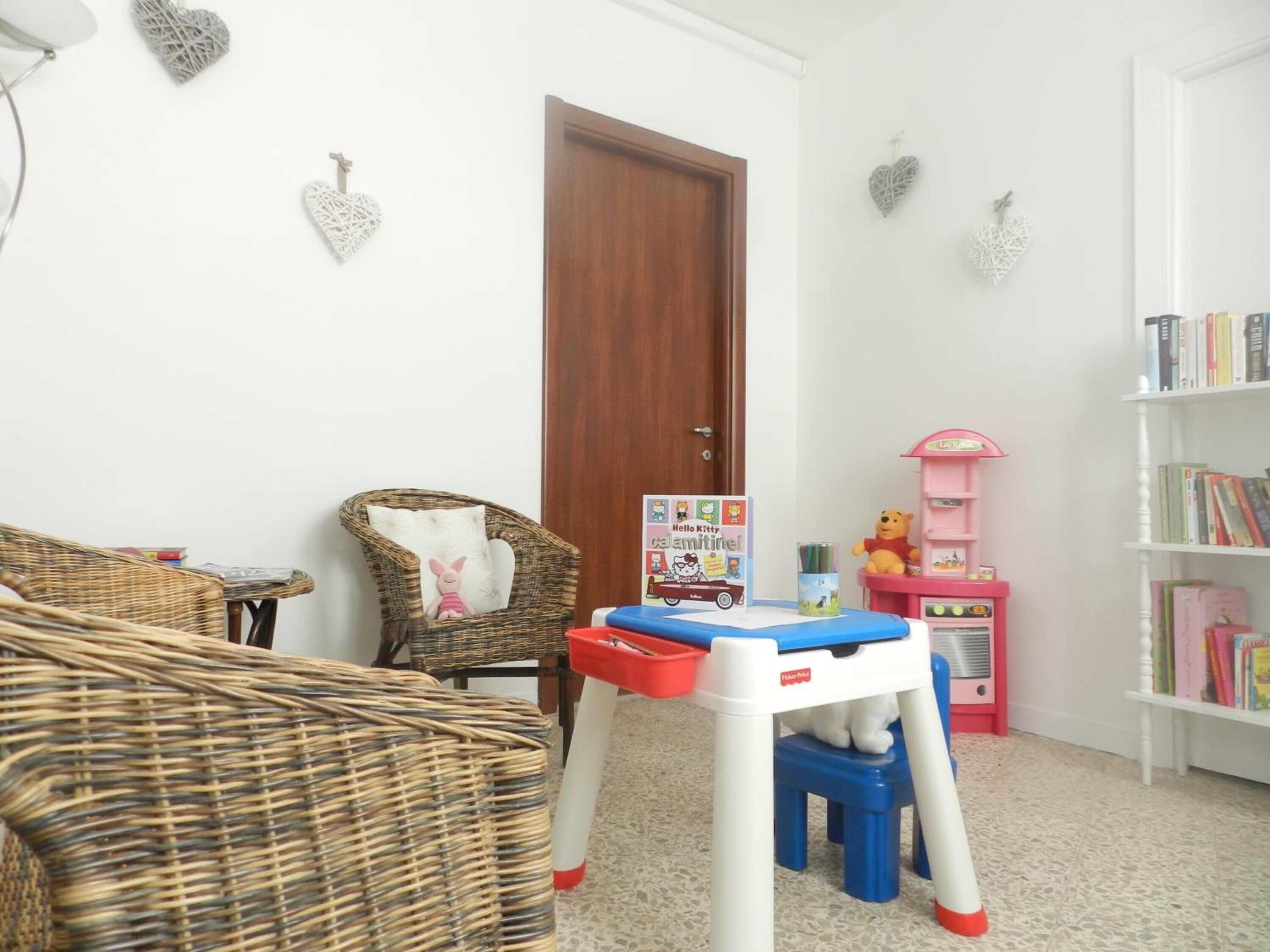 A cozy living room with a wicker sofa, a small table with toys, a bookshelf, and a door.