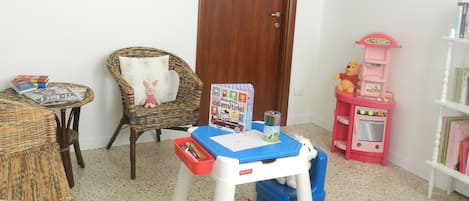 A cozy room with a wicker chair, a small table with books and a stuffed toy, a door, and a bookshelf with children's books and toys.