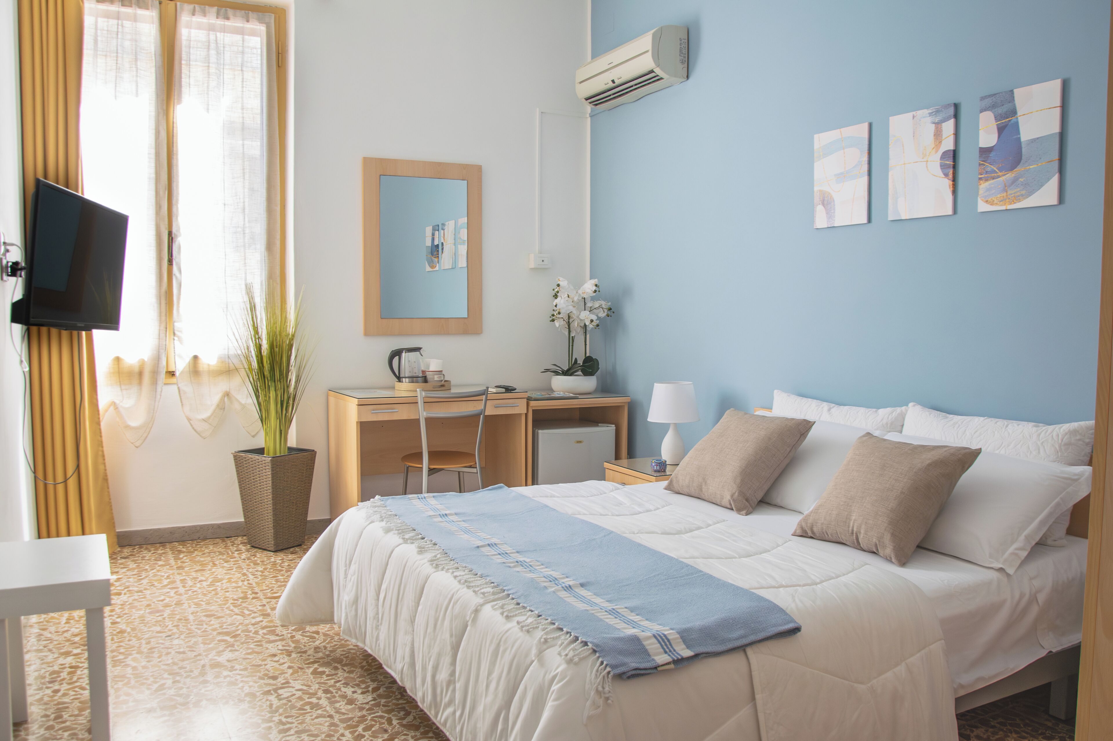Una habitación de hotel con cama, escritorio, silla, televisor, aire acondicionado, una obra de arte y una planta en maceta.