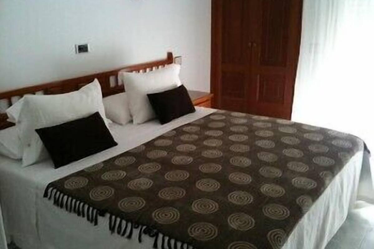 A neatly made bed with a patterned bedspread and pillows, a wooden headboard, and a wooden door in the background.