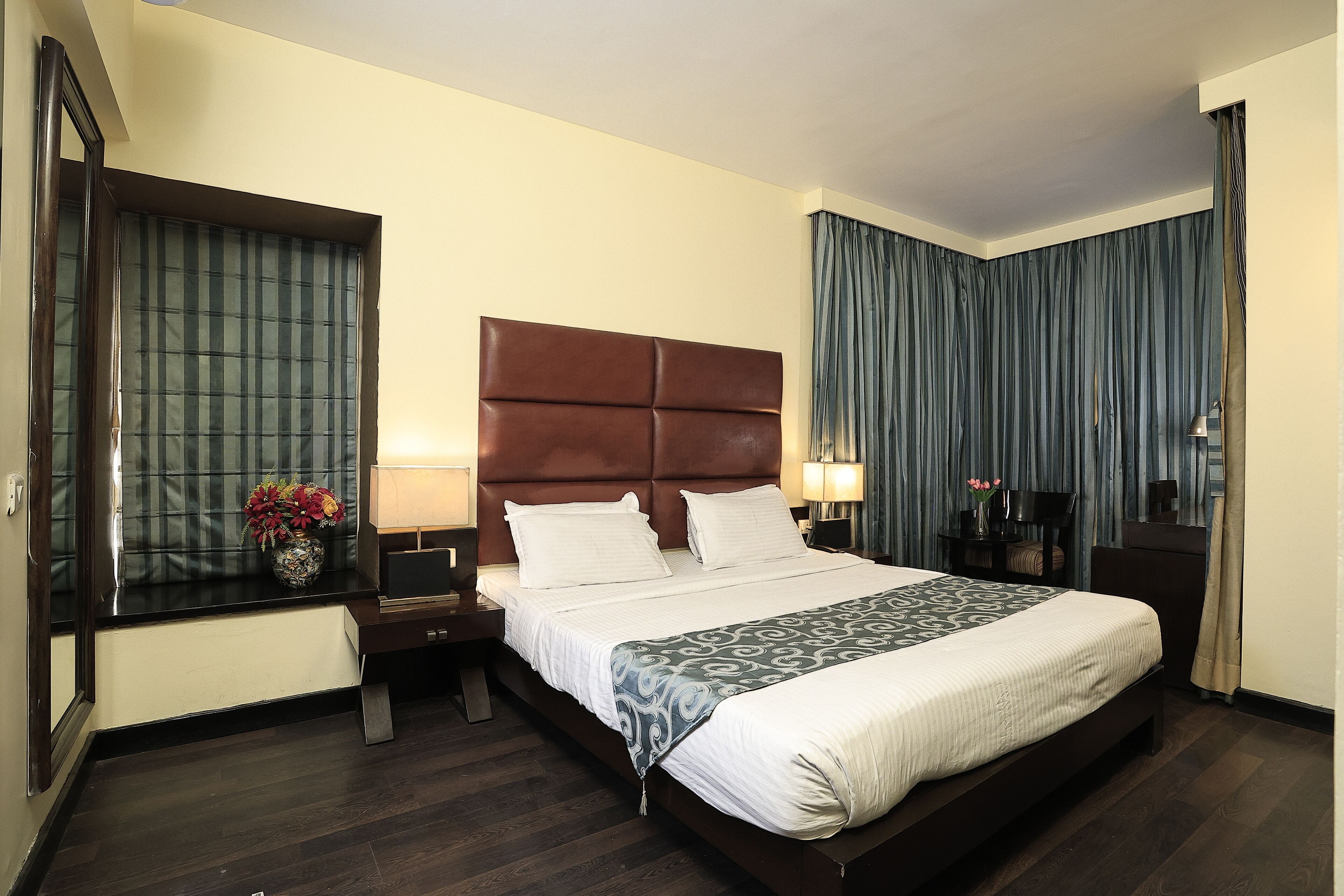 A hotel room with a large bed, a nightstand, a lamp, a vase with flowers, and a window with curtains.