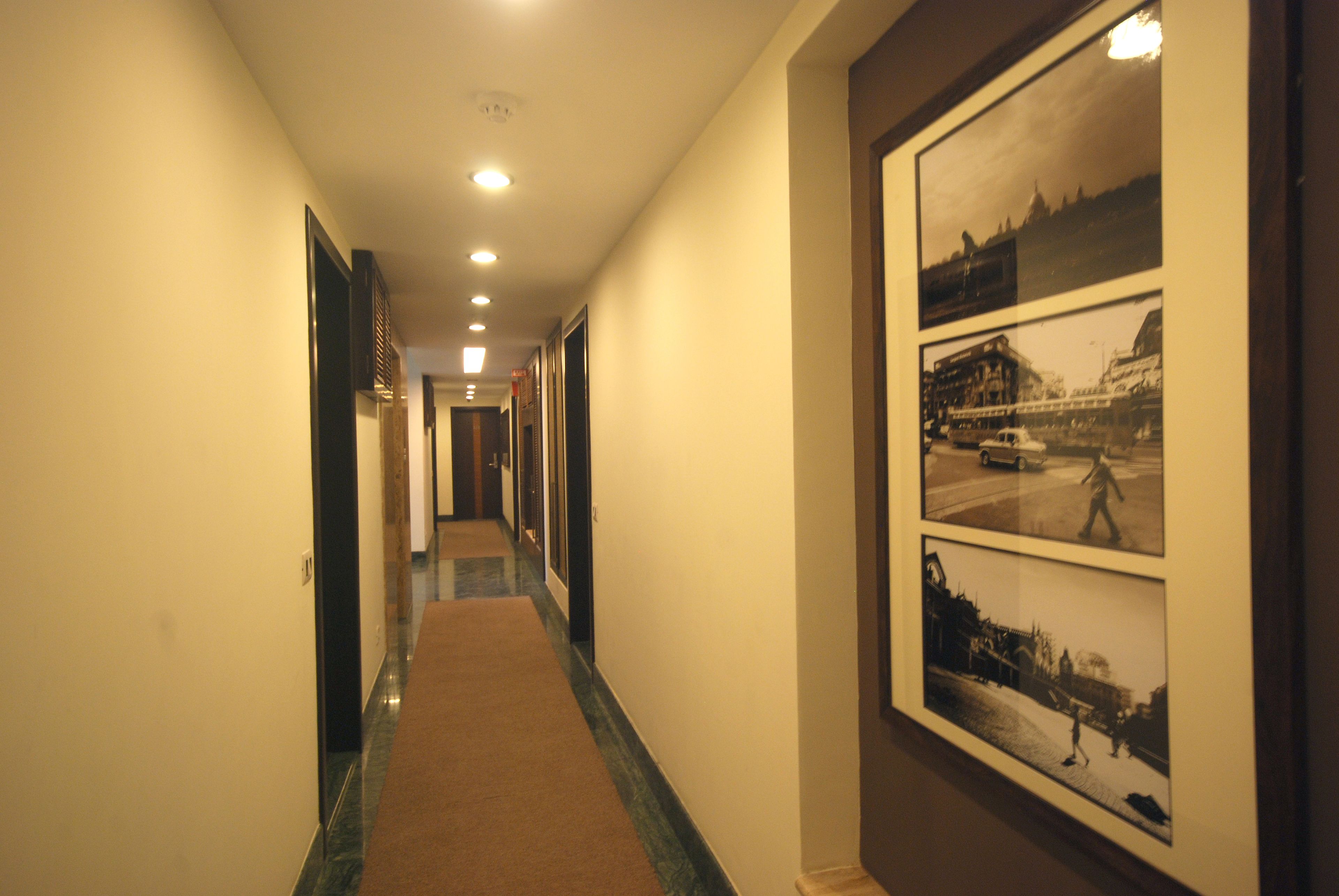A hotel hallway with a carpeted floor, framed pictures on the wall, and recessed lighting.