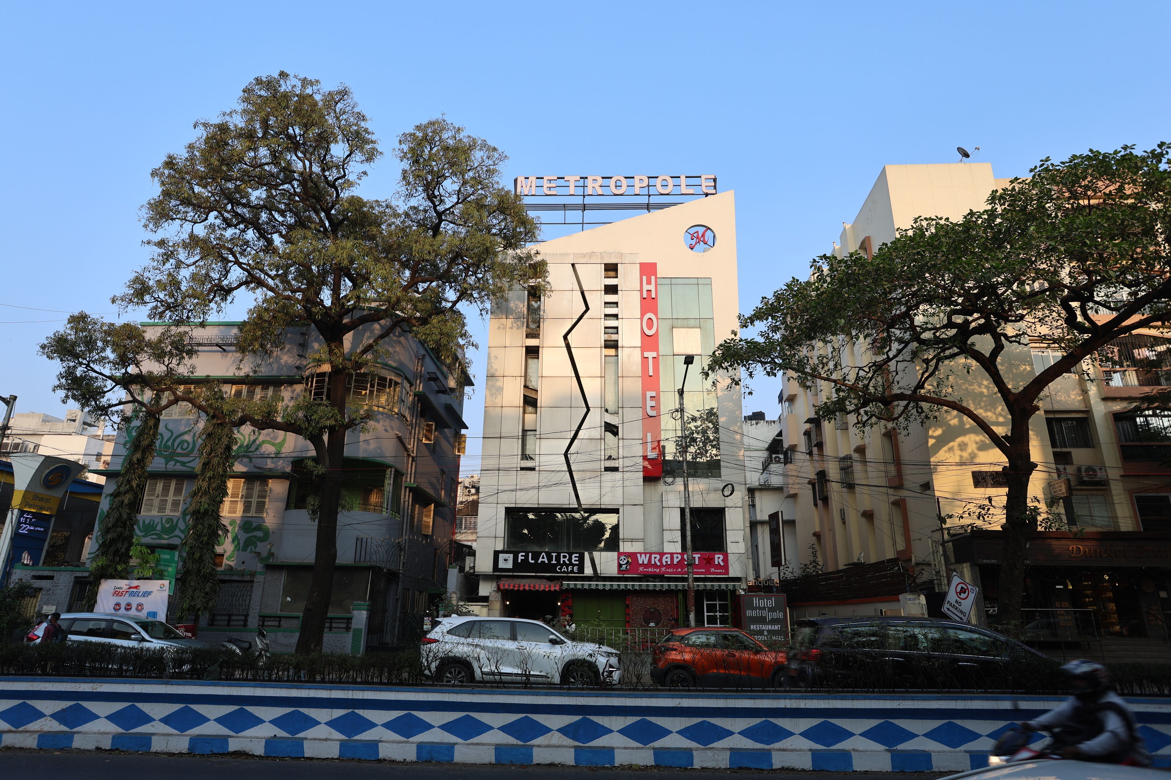 Um hotel chamado "METROPOLIS HOTEL" com uma placa, um café chamado "FLAIRE CAFE" abaixo dela e um restaurante chamado "WRAPSTER" do outro lado da rua.