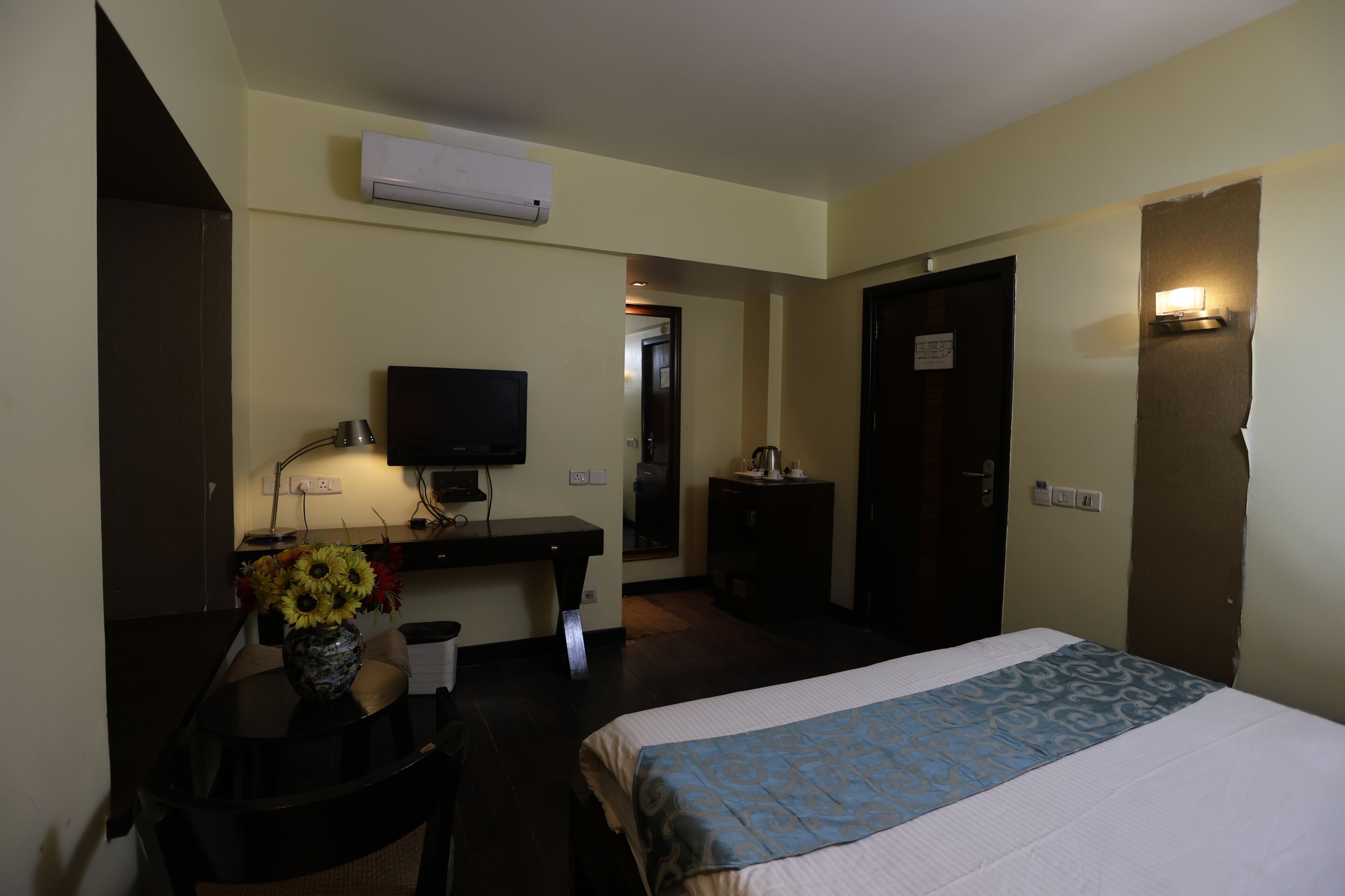 A hotel room with a bed, a desk with a TV, a vase with flowers, and an air conditioning unit.