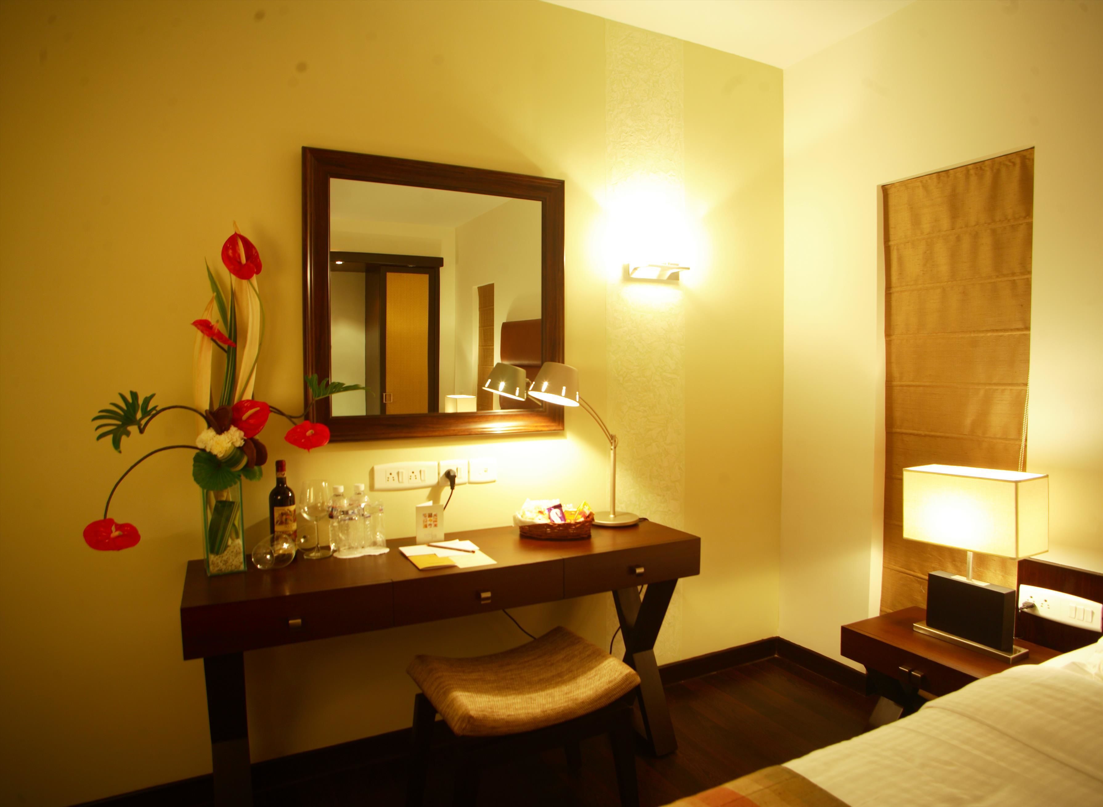 A hotel room with a wooden desk, a mirror, a lamp, a bedside table with a TV, and a bed.
