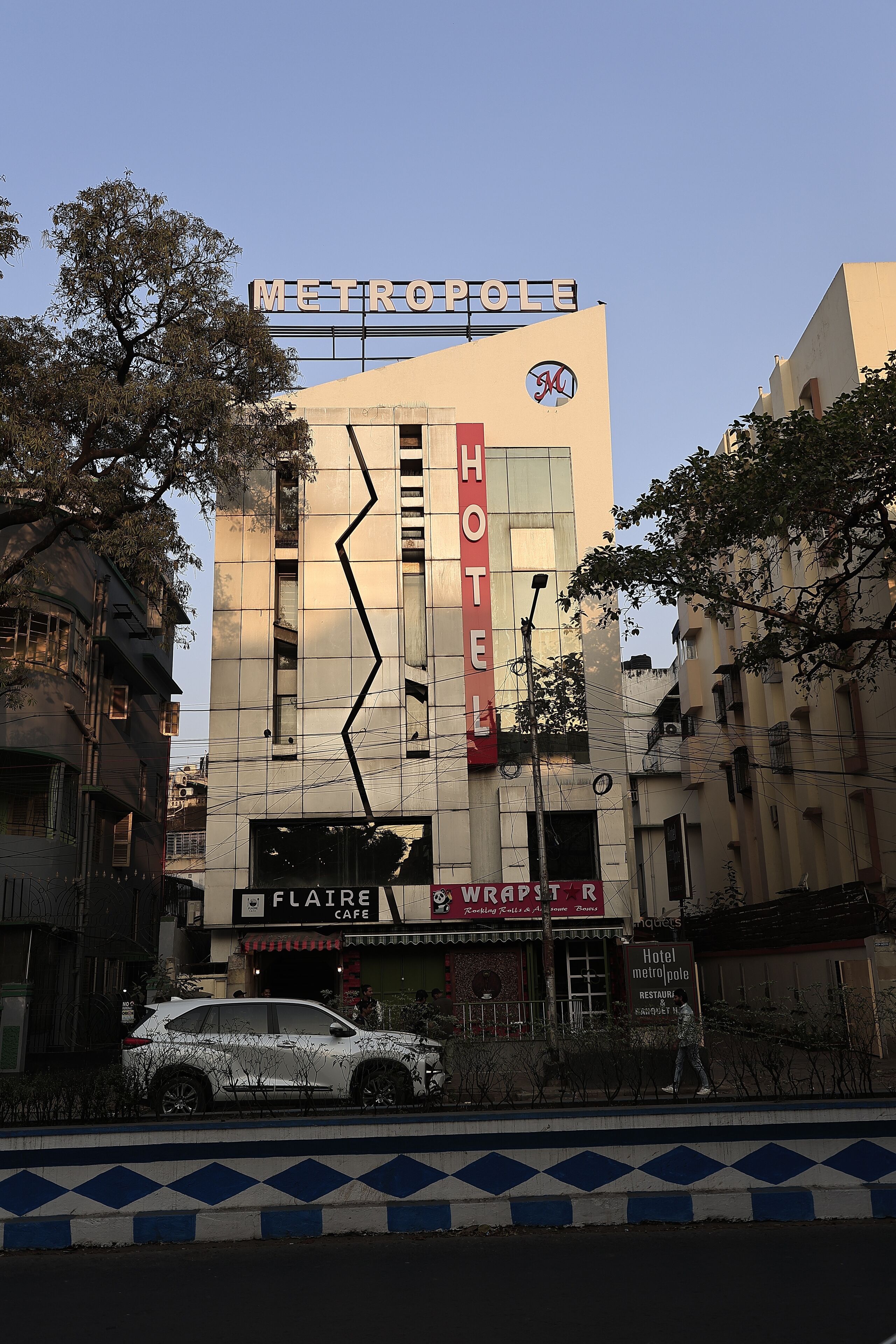 Un hôtel appelé METROPOLI HOTEL, doté d’une façade moderne, d’une entrée bien visible et d’une enseigne indiquant la présence d’un café nommé FLAIRE.