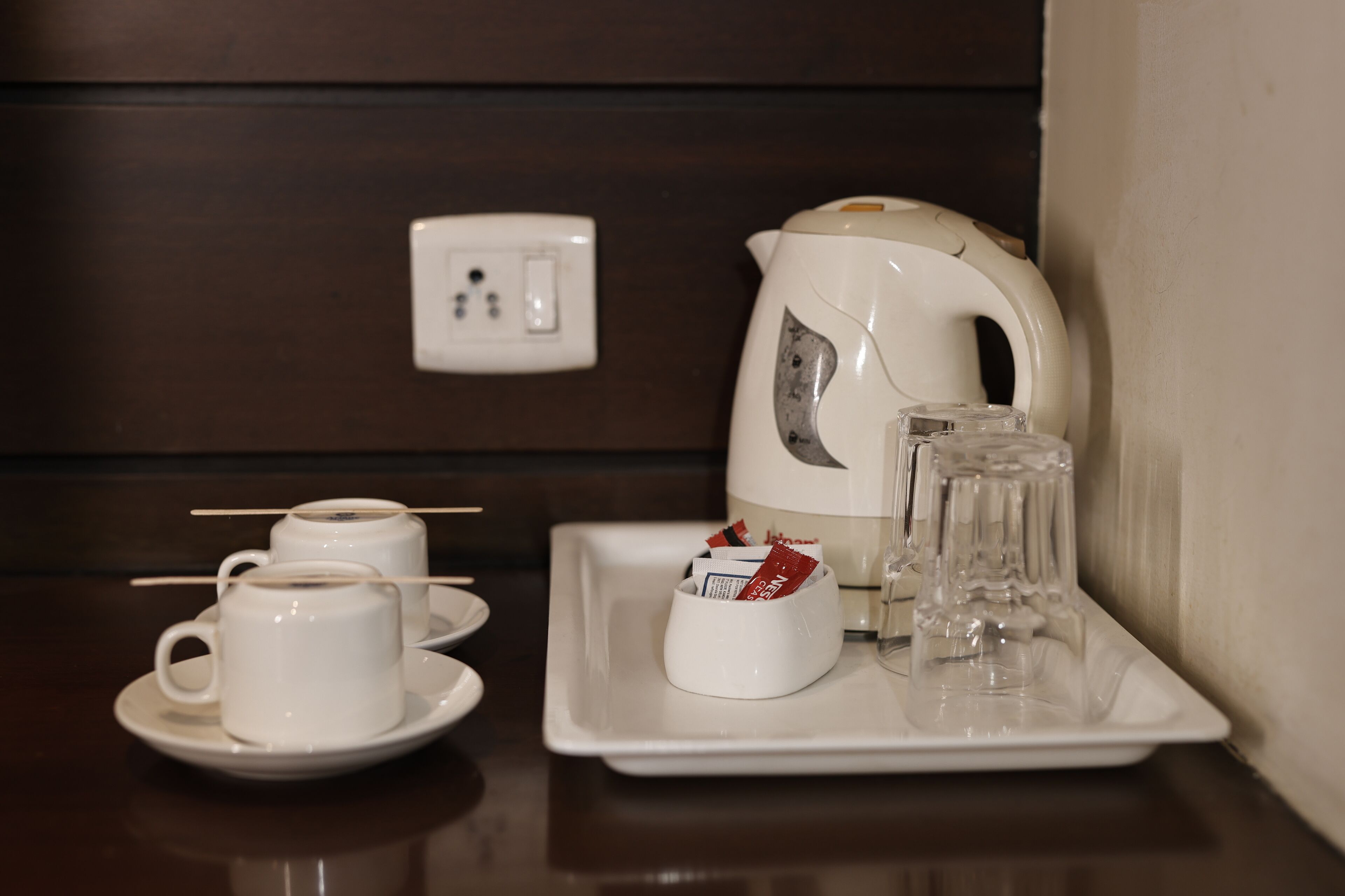 A tray with a white electric kettle, two white cups with straws, a glass, and a small white bowl containing packets.