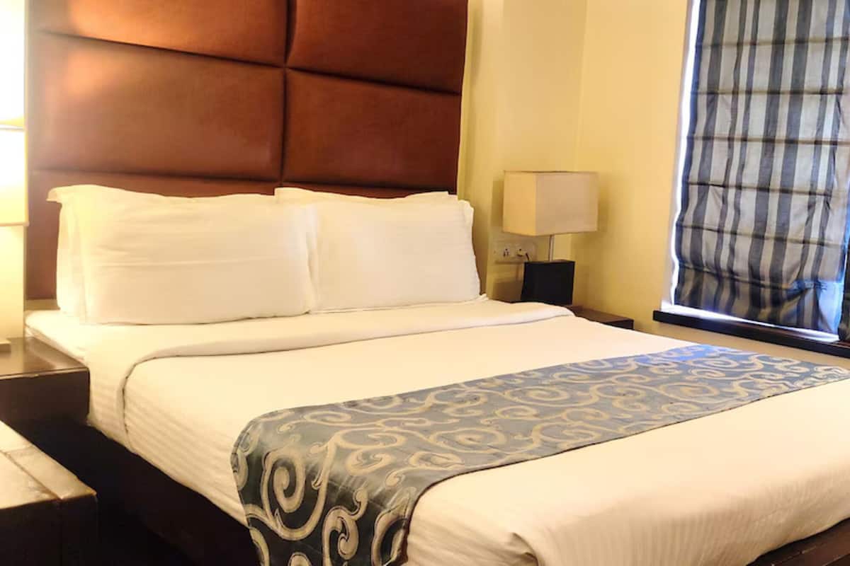 Habitación de hotel con una cama grande, cabecera, mesita de noche, lámpara y ventana con cortinas.