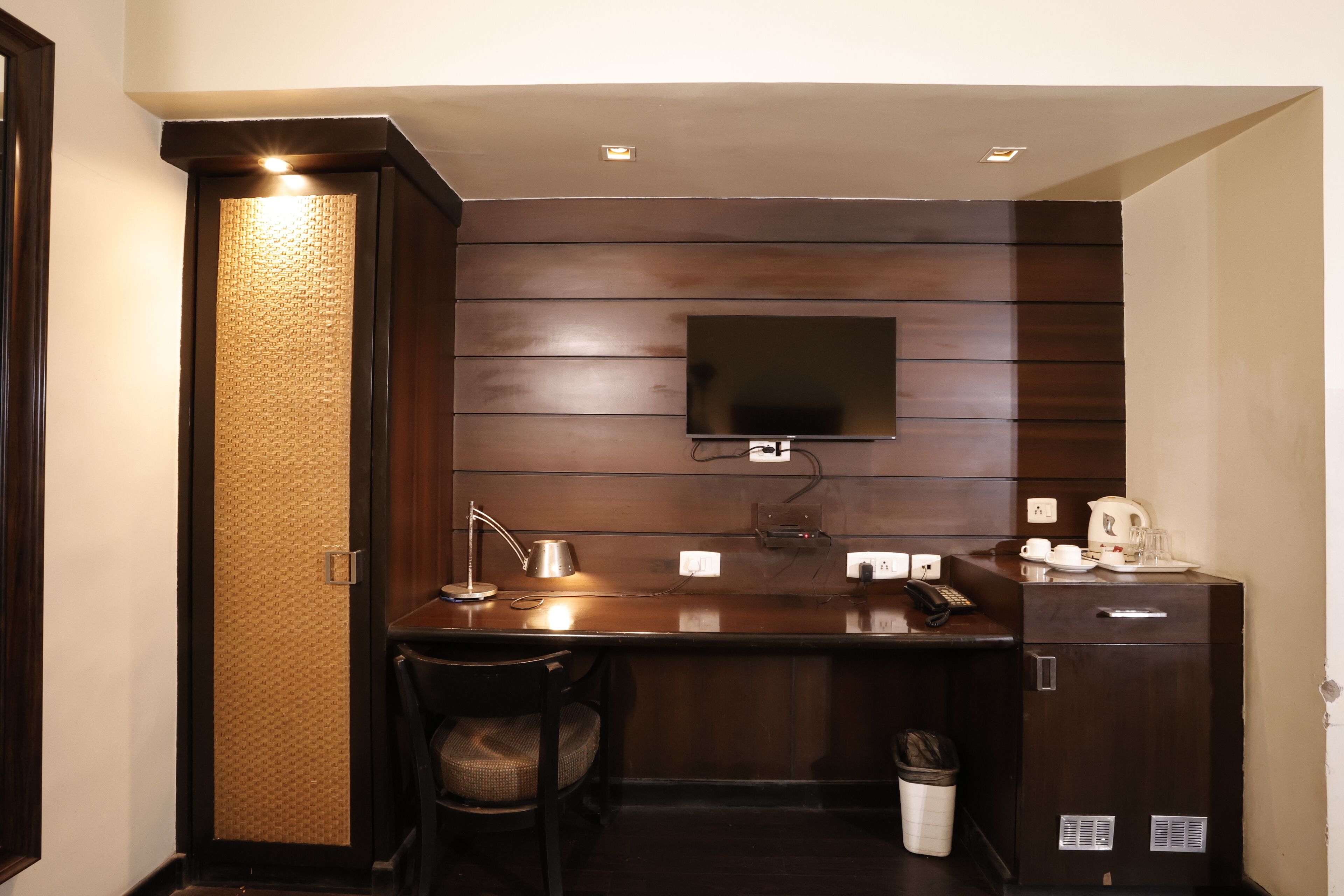 A compact hotel room with a dark wood panel wall, a flat-screen TV mounted above a wooden desk, a chair, a small trash bin, and a cabinet with a kettle and cups.