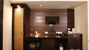A compact hotel room with a dark wood panel wall, a flat-screen TV mounted above a wooden desk, a chair, a small trash bin, and a cabinet with a kettle and cups.