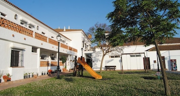 Un patio con un parque infantil, que incluye un tobogán, rodeado de edificios blancos con detalles en rojo y un árbol.