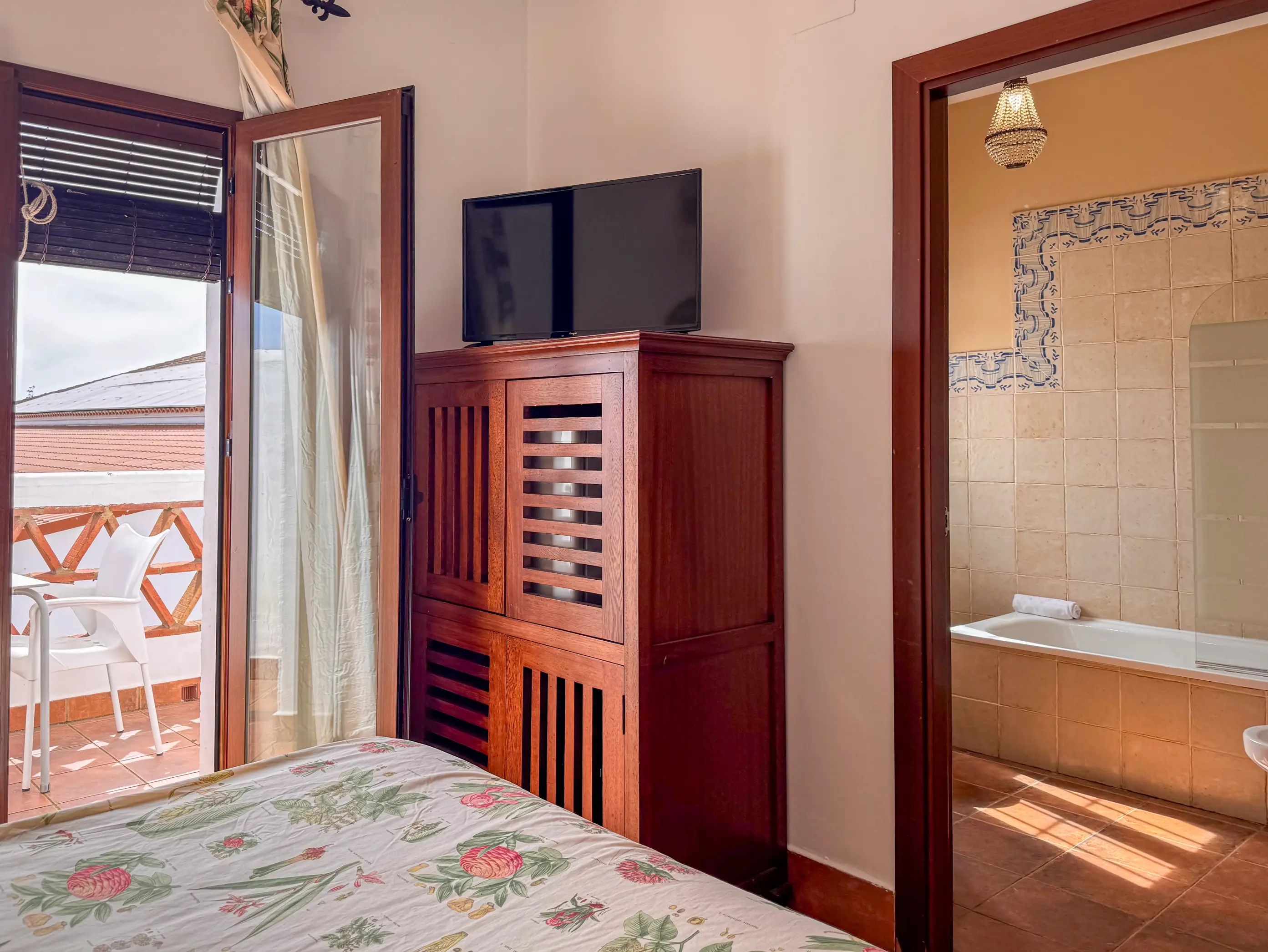 Een slaapkamer met een bed, een houten garderobe, een televisie erop, een badkamer met een bad en uitzicht op het balkon.