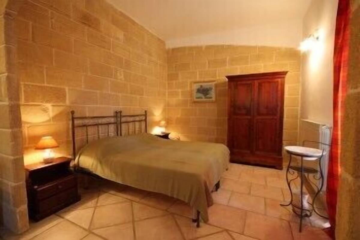 A bedroom with a bed, a nightstand, a wooden wardrobe, a chair, and a wall-mounted light fixture.