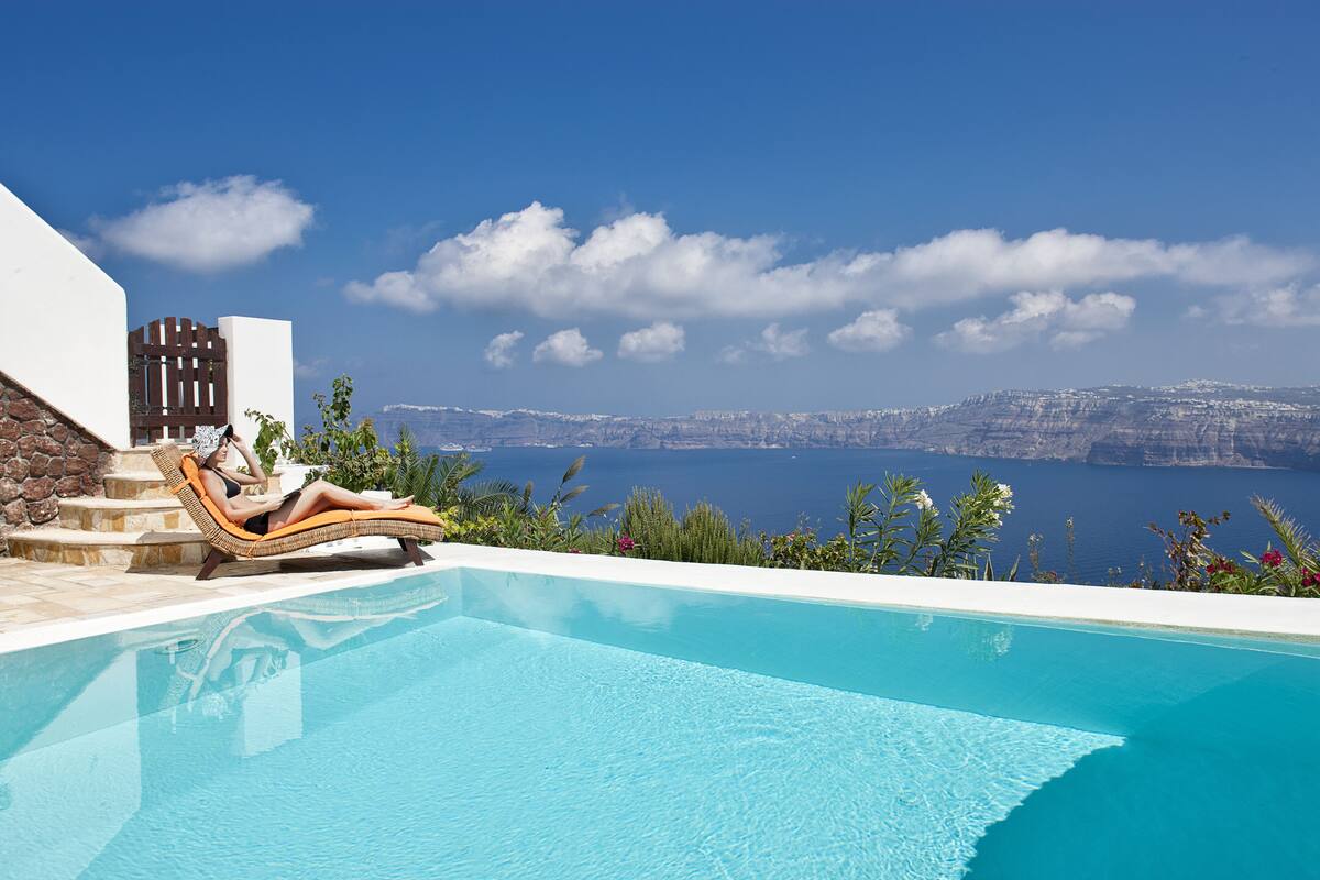 A pool with a person lounging on a chair, a stone wall, and a view of the sea and cliffs.