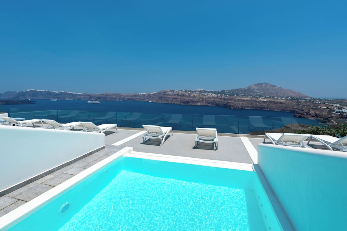 A rooftop pool with clear blue water, surrounded by white lounge chairs, offering a view of the sea and distant mountains.